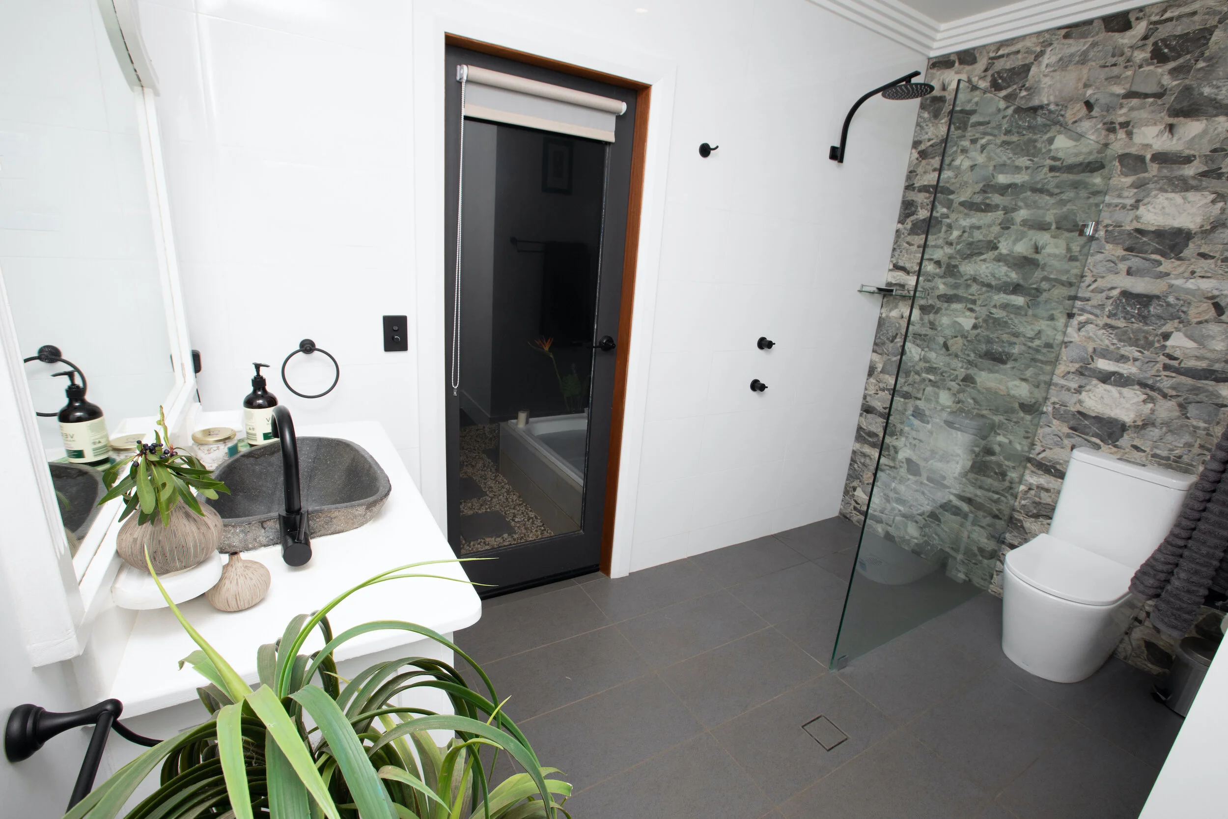 Modern bathroom with grey tiled floor, stone accent wall shower area with black showerhead, white toilet, black sink with a plant on the countertop, and a black door leading to a dark shower area, decorated with black fixtures and white walls.