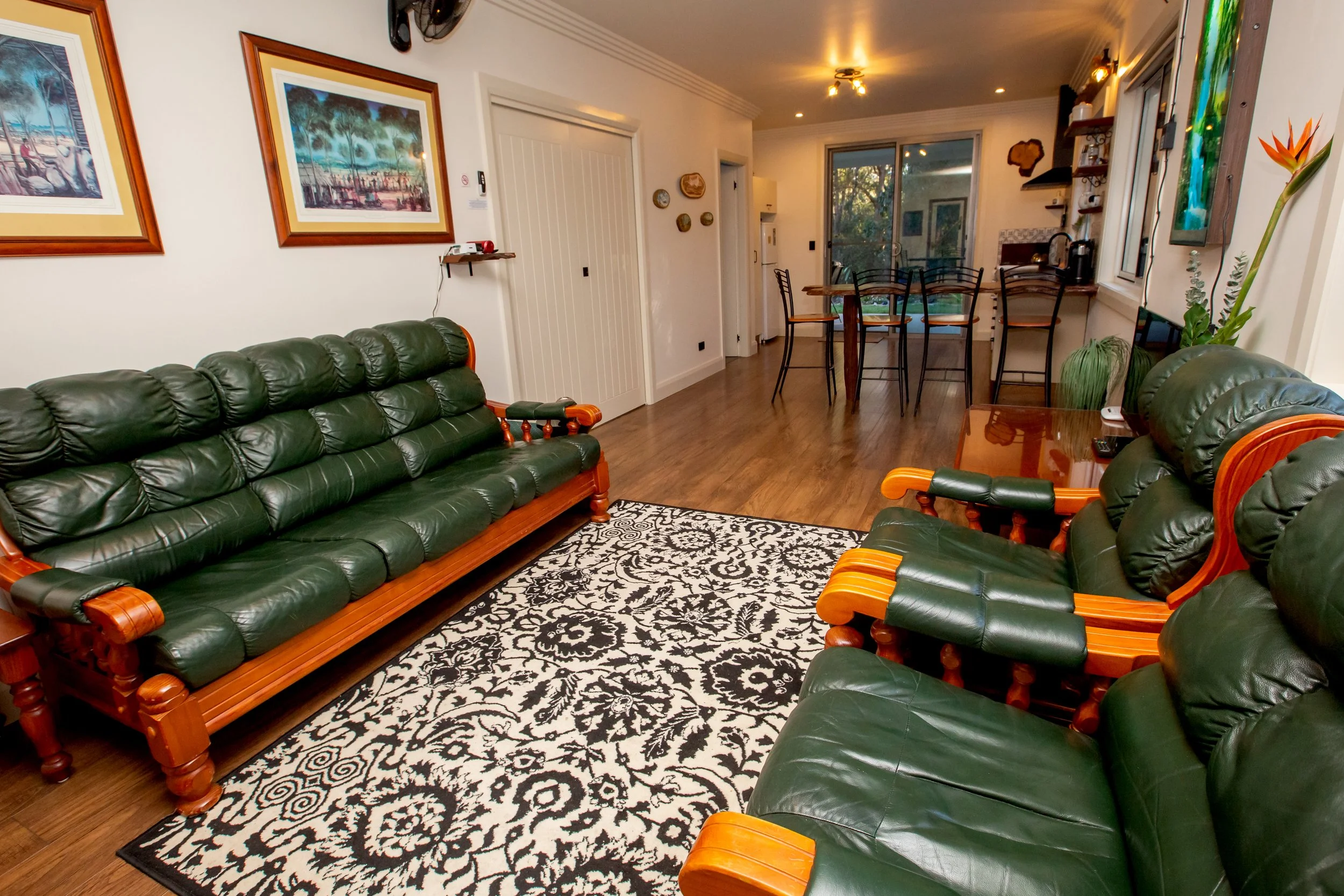 Living room with green leather sofas, a black and white patterned rug, wall art, a kitchen table with chairs, and a sliding glass door leading outside.