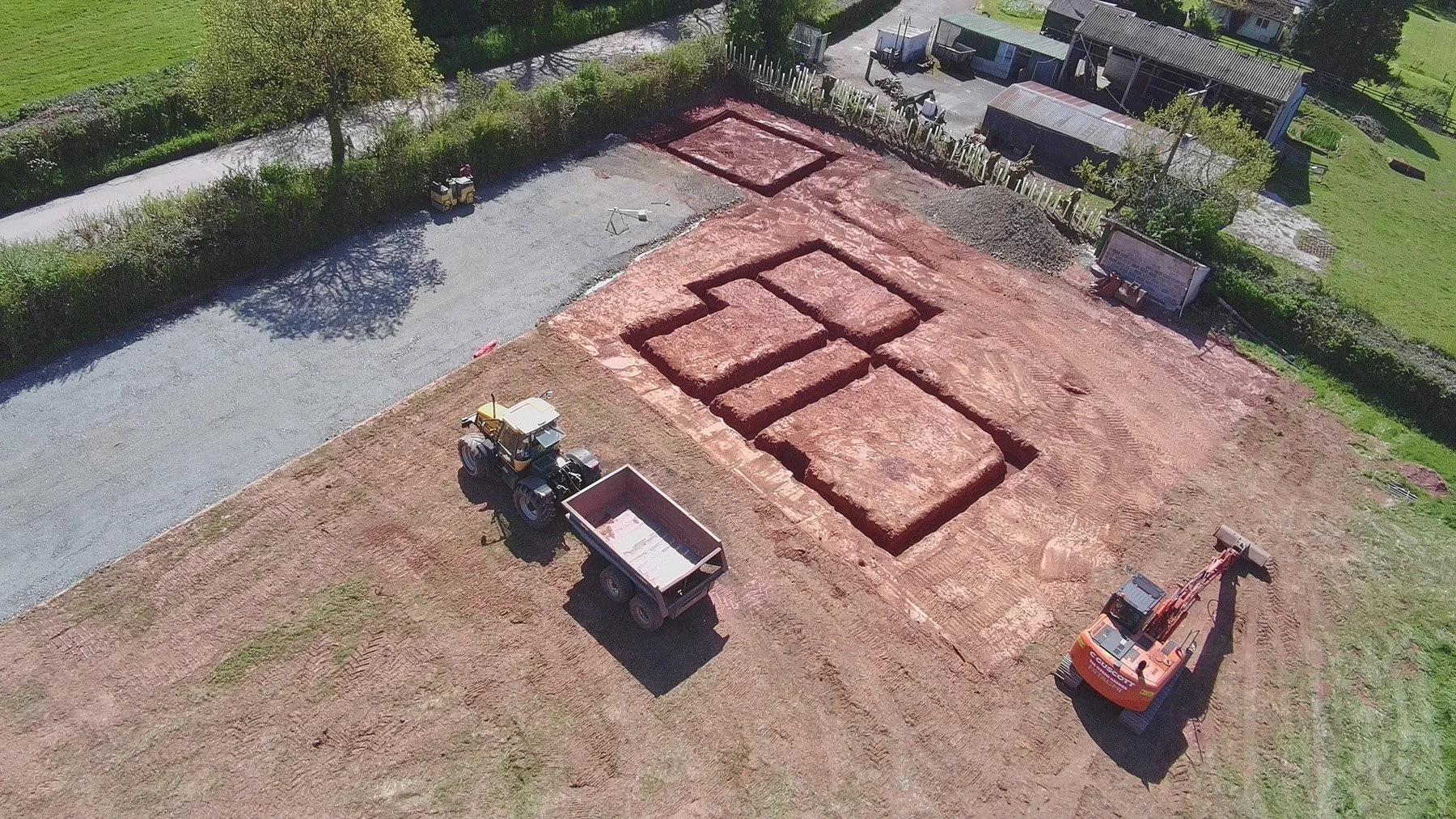 An aerial view of a construction site showing the initial stage of foundation work with holes dug in the ground, construction machinery, and surrounding greenery.