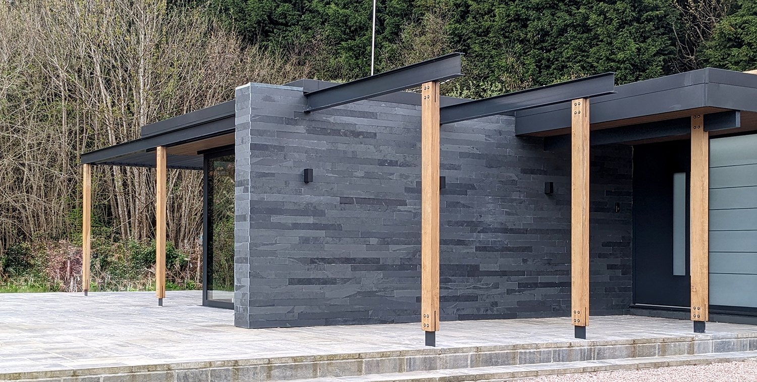 Contemporary bungalow extension at Park Corner, Devon, featuring natural slate cladding, exposed timber posts, an integrated pergola and a landscaped terrace.