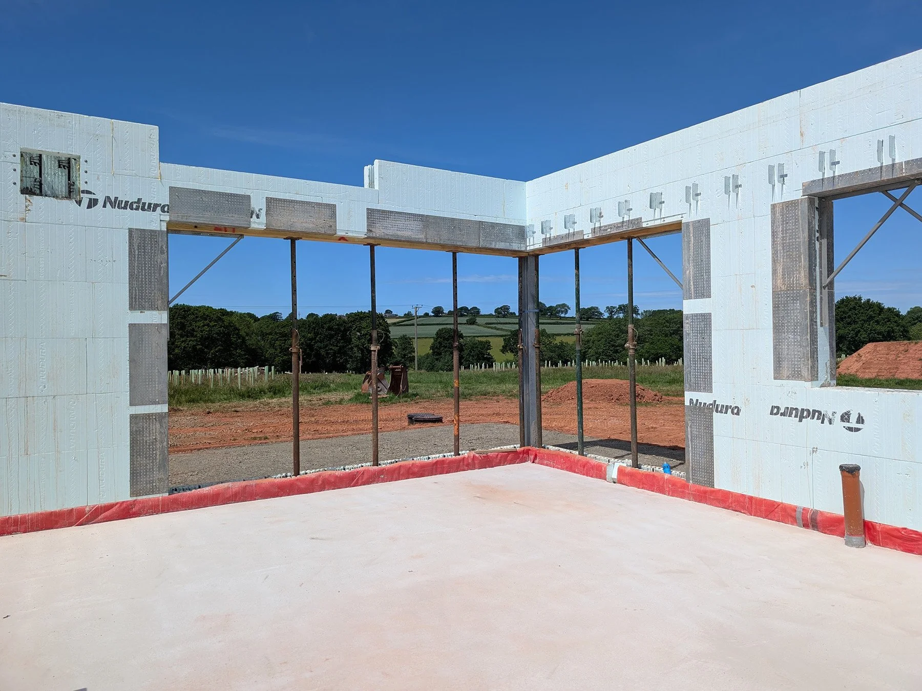 Construction site with the framed structure of a building under development, featuring insulation and support beams, with clear skies and distant fields in the background.