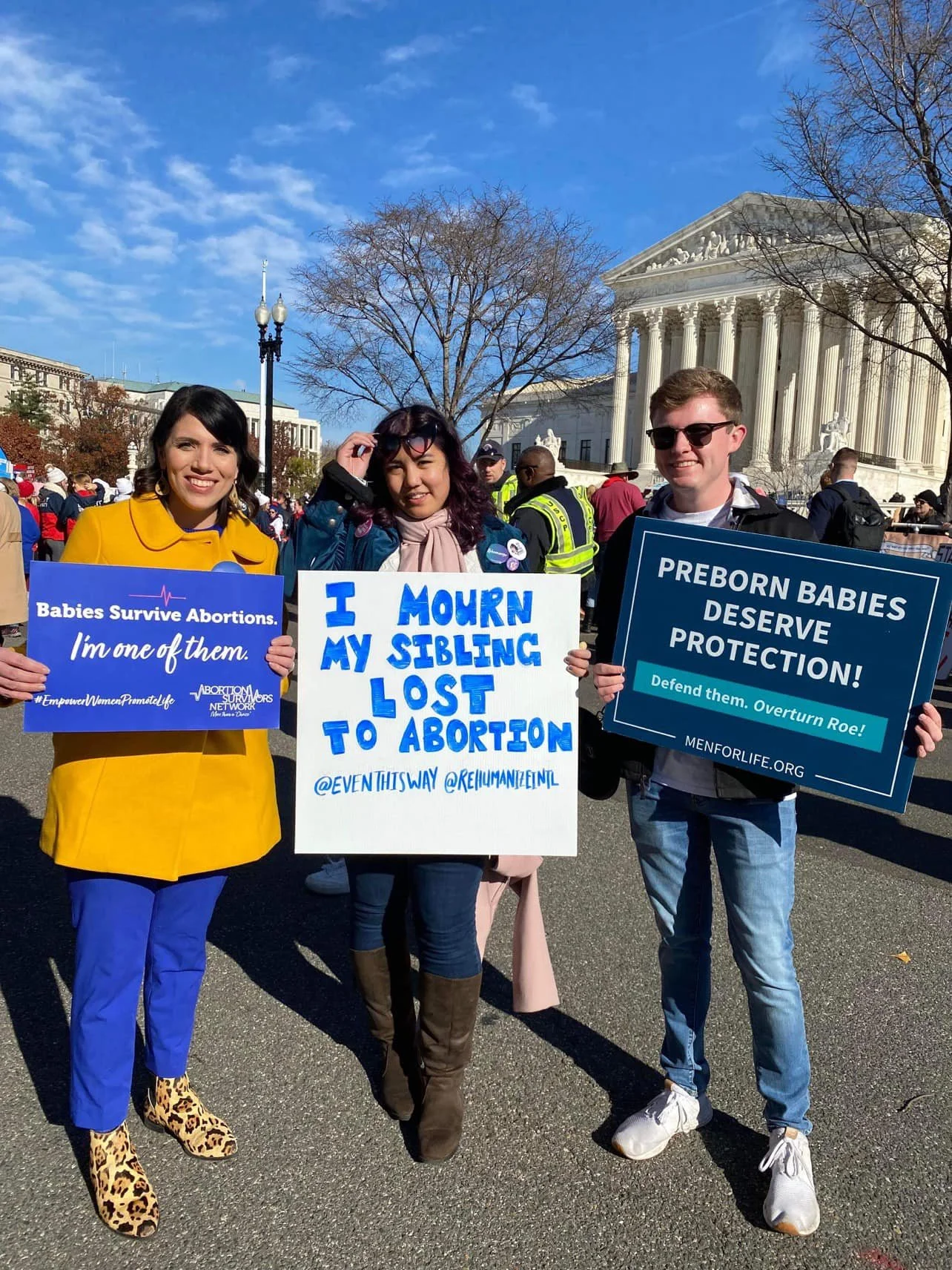 Three people standing outdoors at a protest, holding signs supporting abortion rights, with the U.S. Supreme Court building in the background.