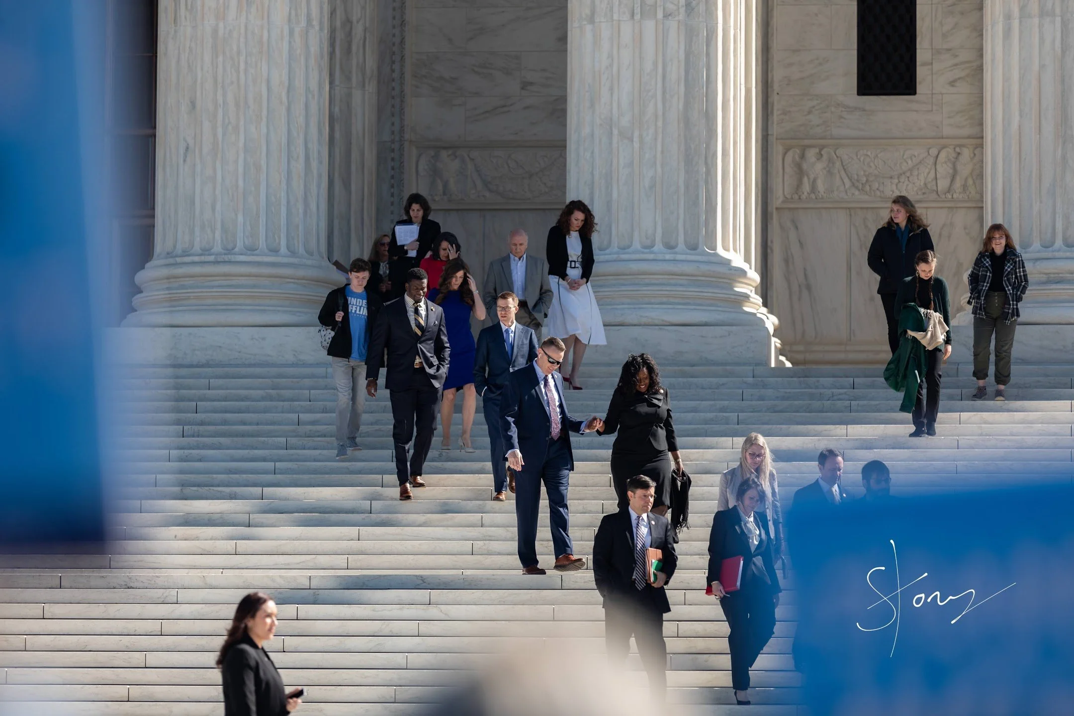 People descending the steps of a government building with large marble columns, engaged in conversation or focused, some holding documents or phones.