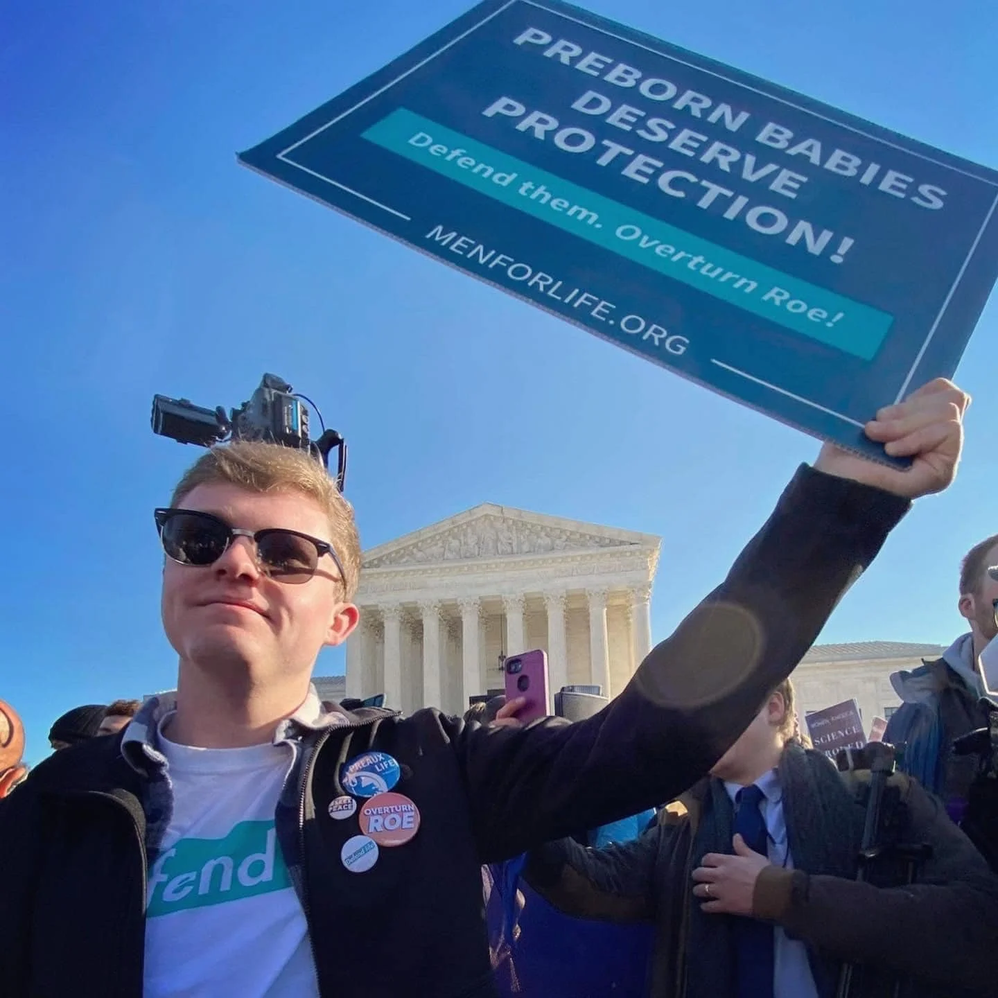 A young man wearing sunglasses and buttons supporting reproductive rights holds a sign protesting preborn babies' DEPROVE and protect their future, with the Capitol building in the background.