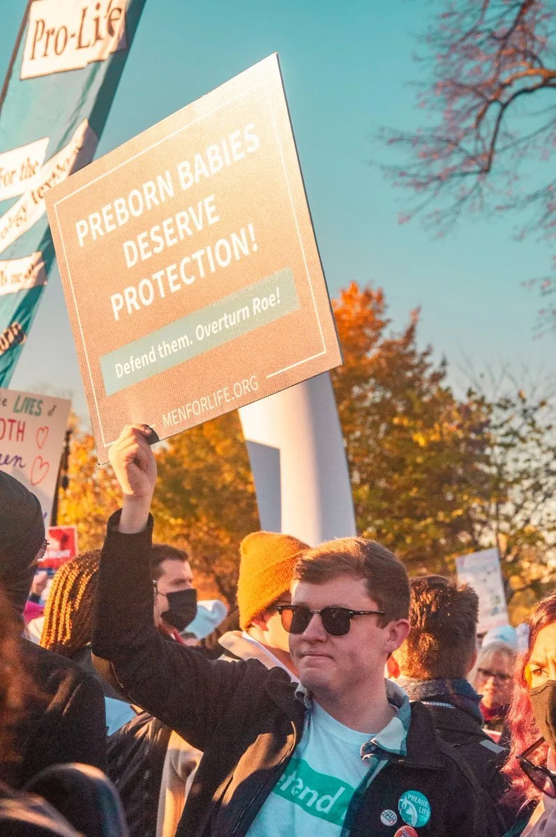 A young man wearing sunglasses and a black jacket holding a sign at a protest. The sign reads, "Preborn Babies Deserve Protection! Defend them. Overturn Roe!" with the website menforlife.org at the bottom. Other protesters and autumn trees are visible in the background.