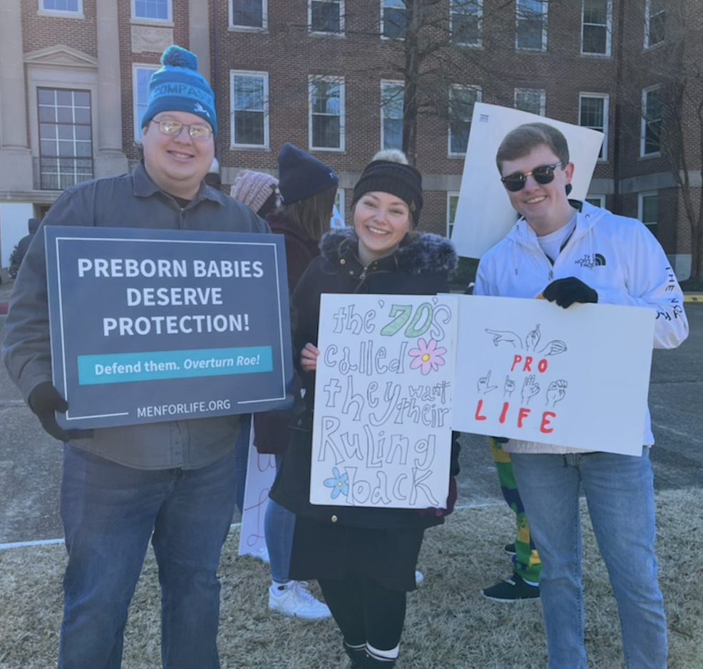 Three young adults standing outdoors at a protest, holding signs supporting preborn babies and pro-life causes. The person on the left has a blue beanie and glasses, holding a sign that reads 'Preborn Babies Deserve Protection!'. The middle person is smiling and holding a sign that says 'the '70s called, they want their ruling back' with colorful lettering and drawings. The person on the right is wearing sunglasses and a white jacket, holding a sign with messages about 'PRO LIFE' and illustrations of hand gestures.