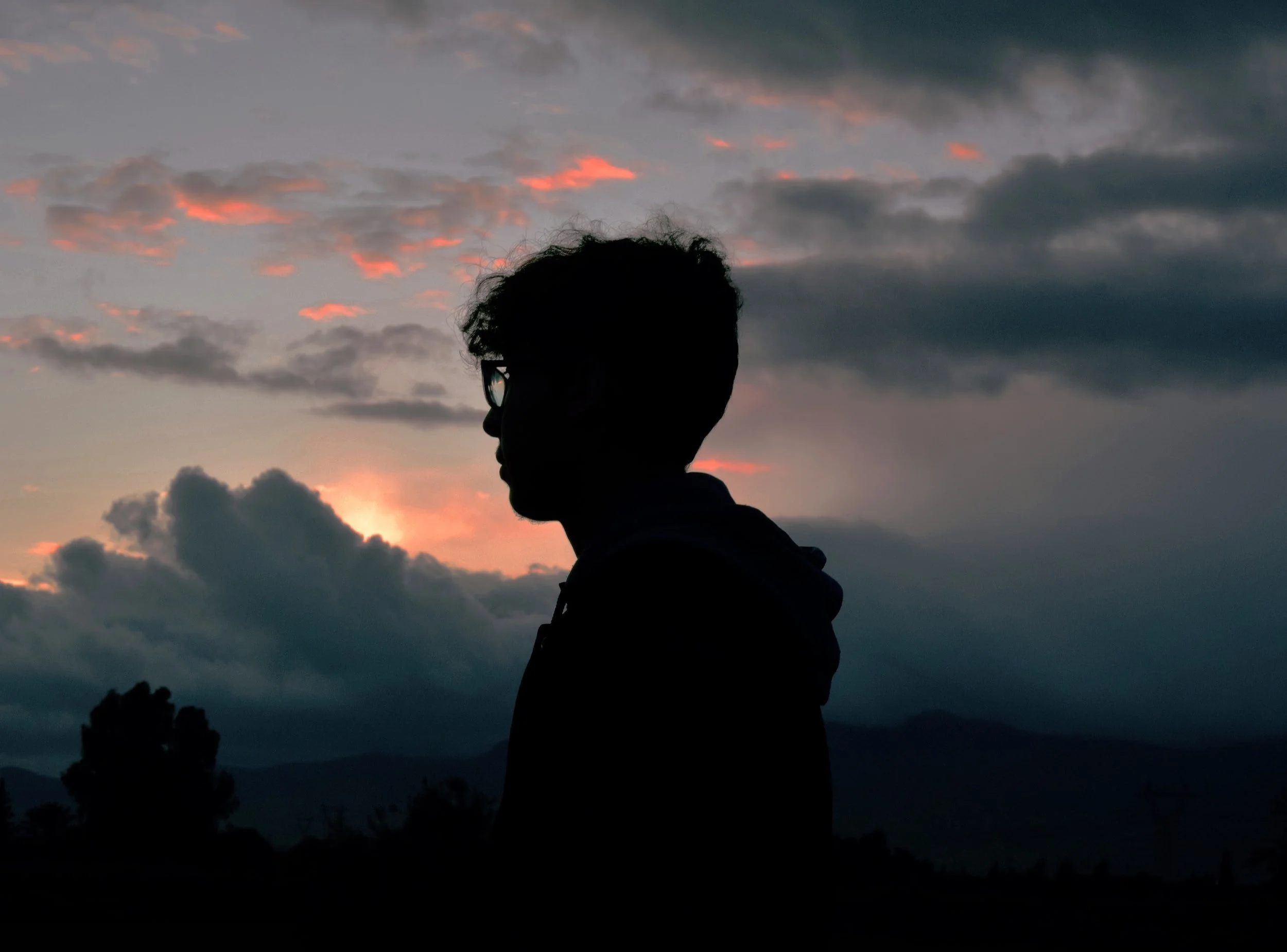 Silhouette of a person with glasses standing outdoors during sunset with colorful clouds and distant mountains in the background.