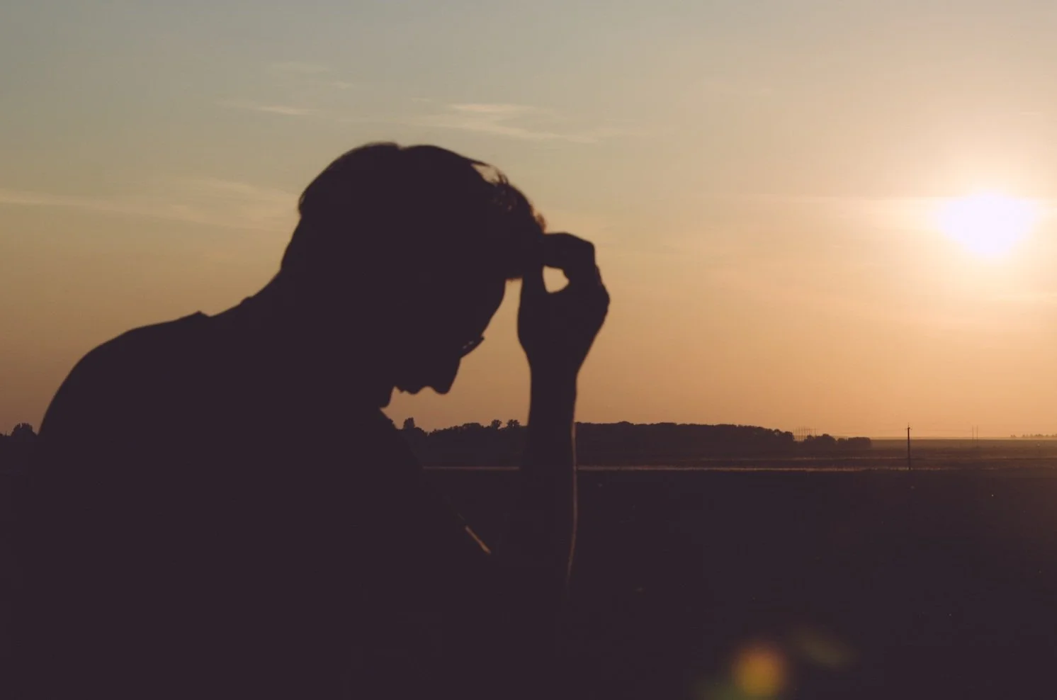 Silhouette of a person with head bowed and hand on forehead, during sunset with open landscape in the background.