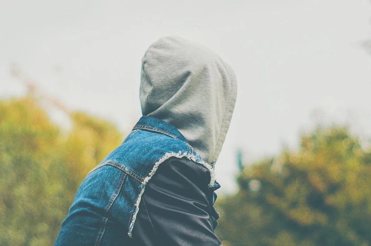 Person wearing a gray hoodie and a denim jacket, facing away outdoors during daytime.