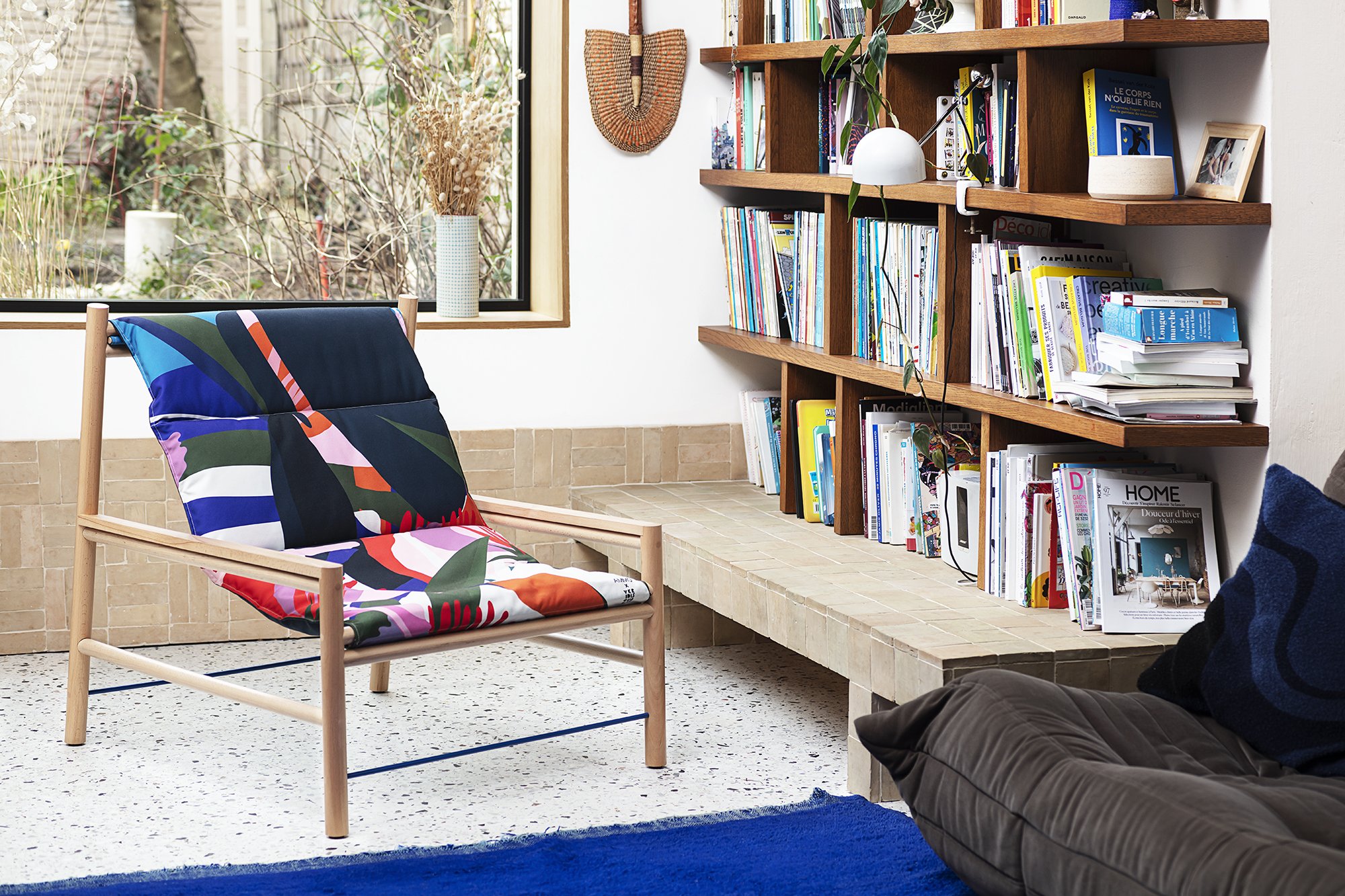Intérieur moderne avec chaise colorée, étagères de livres en bois, fenêtre sur jardin.