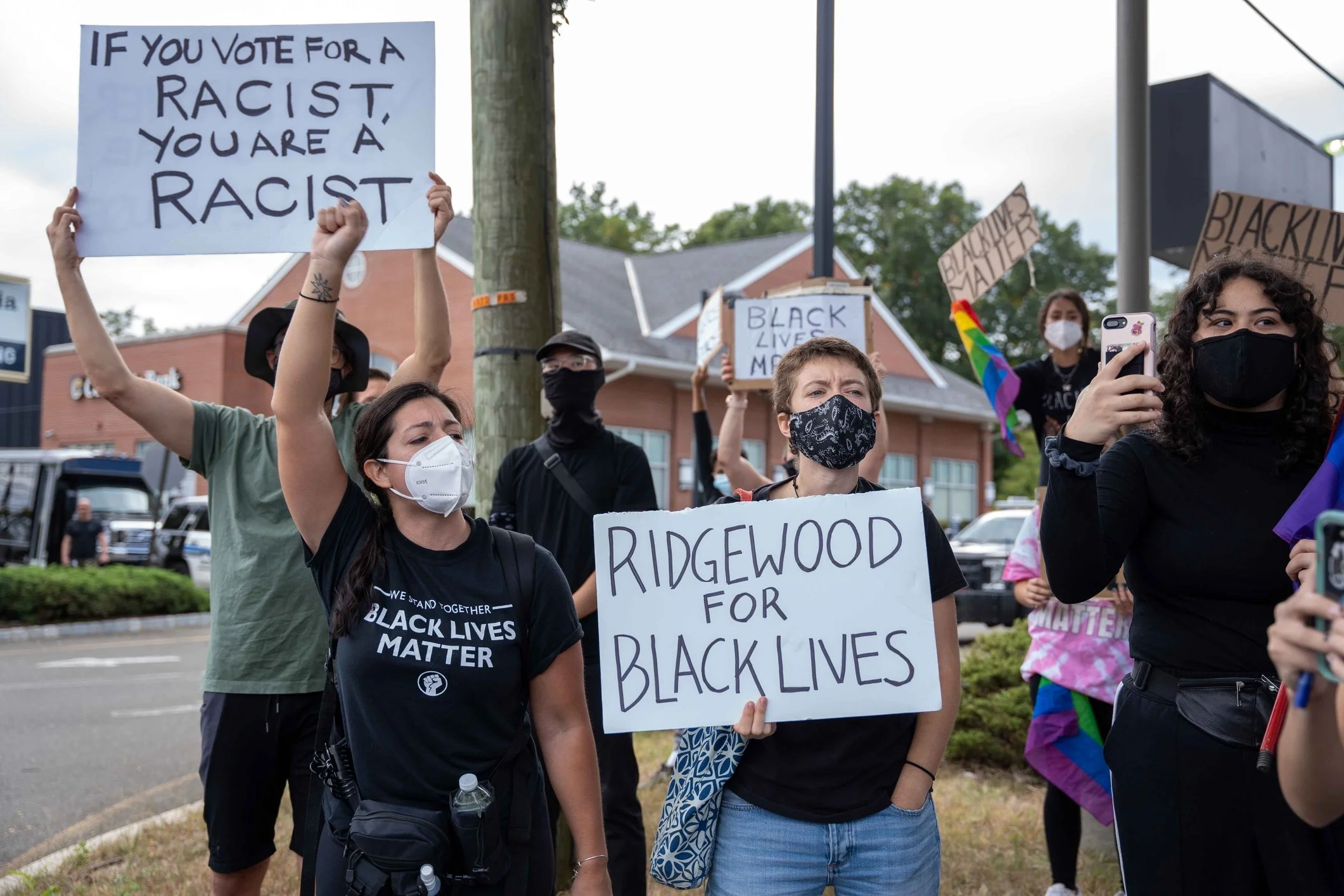 Protesters wearing face masks at a rally holding signs that say 'Ridgewood for Black Lives', 'If you vote for a racist, you are a racist', and 'Black Lives Matter'.