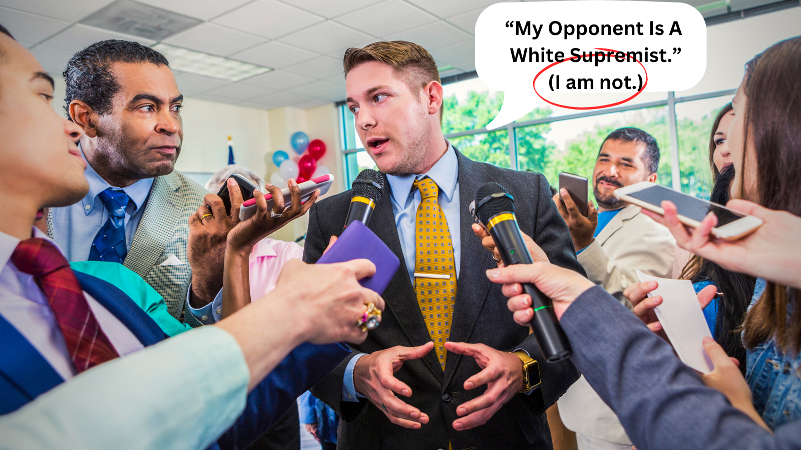 A man in a gray suit and yellow tie is speaking into microphones while surrounded by reporters holding smartphones and recording devices during a press conference or interview in a bright room with balloons in the background.