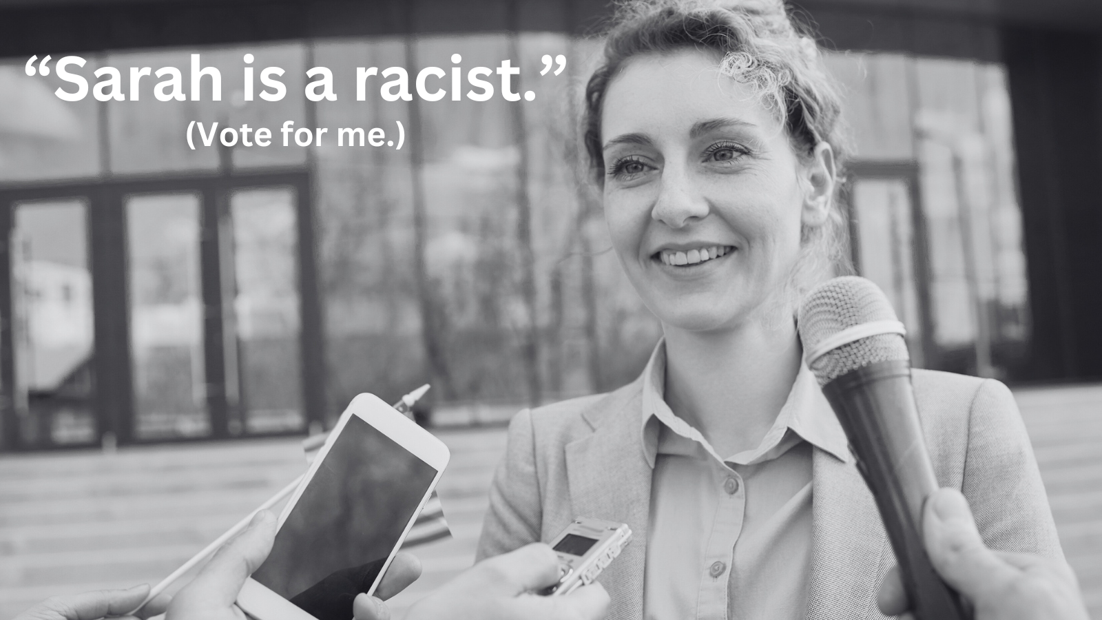 A woman in a blazer smiles while being interviewed with multiple microphones and a smartphone held towards her, outdoors in front of modern glass building.