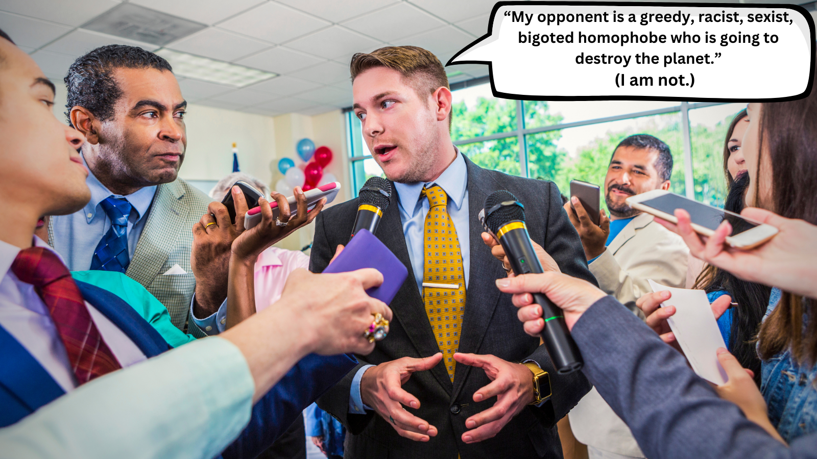 Man in a suit and yellow tie speaking to reporters holding microphones and phones during a press conference, with balloons and windows in the background.