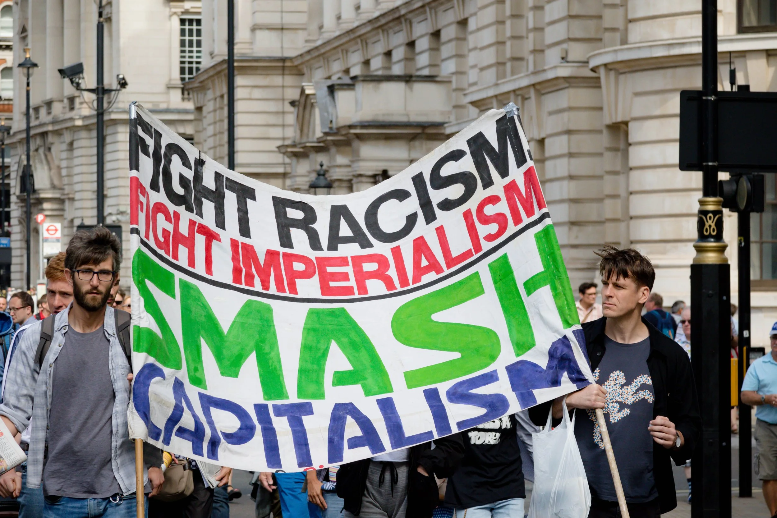 Crowd of people participating in a protest or demonstration holding a large banner with phrases opposing racism and capitalism in an urban city setting.