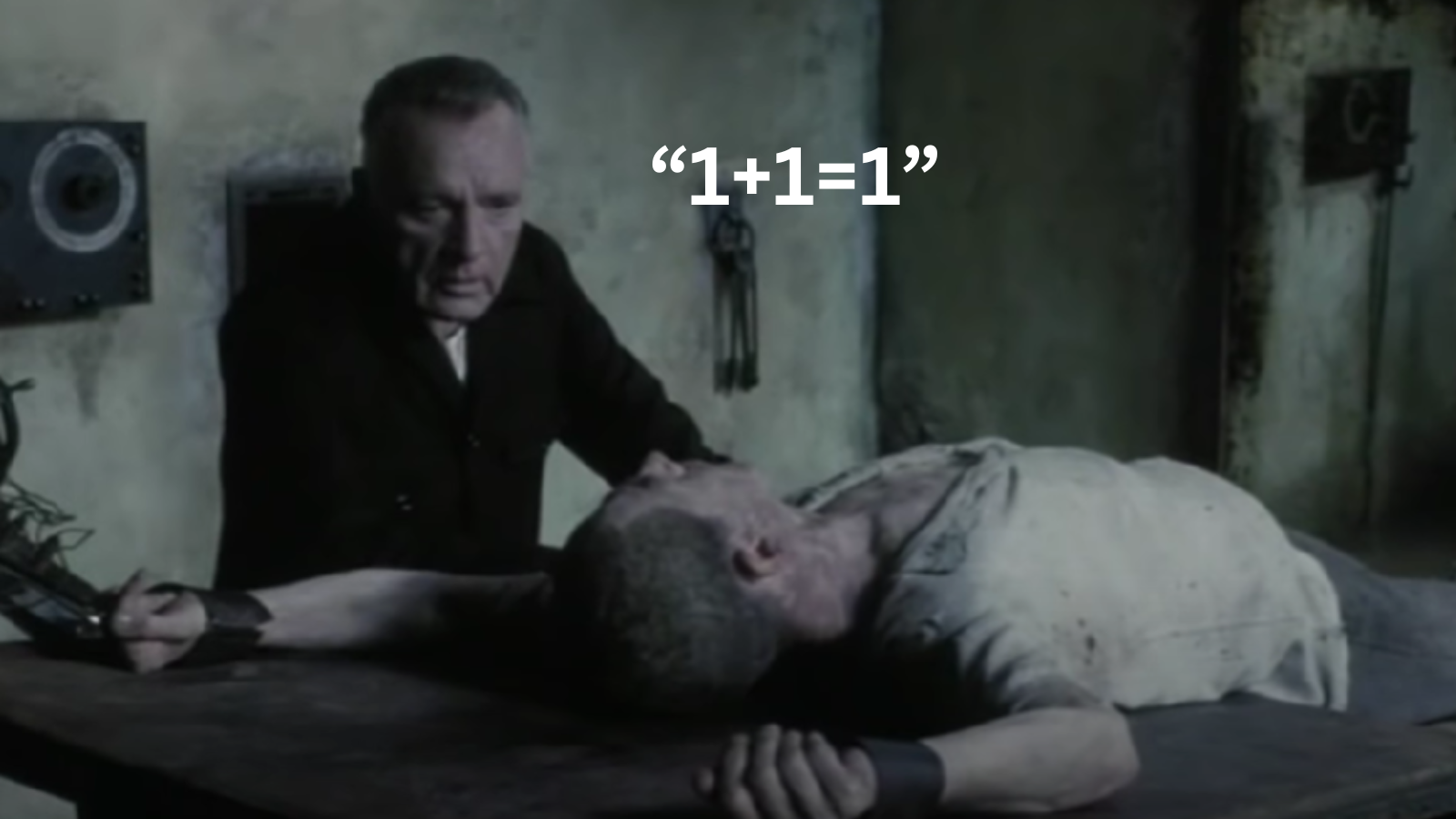A scene from a movie showing a man with a stern expression sitting beside a lifeless body of an older person lying face down on a table. The scene has an old, worn setting with muted colors. Text overlay reads "1+1=1".
