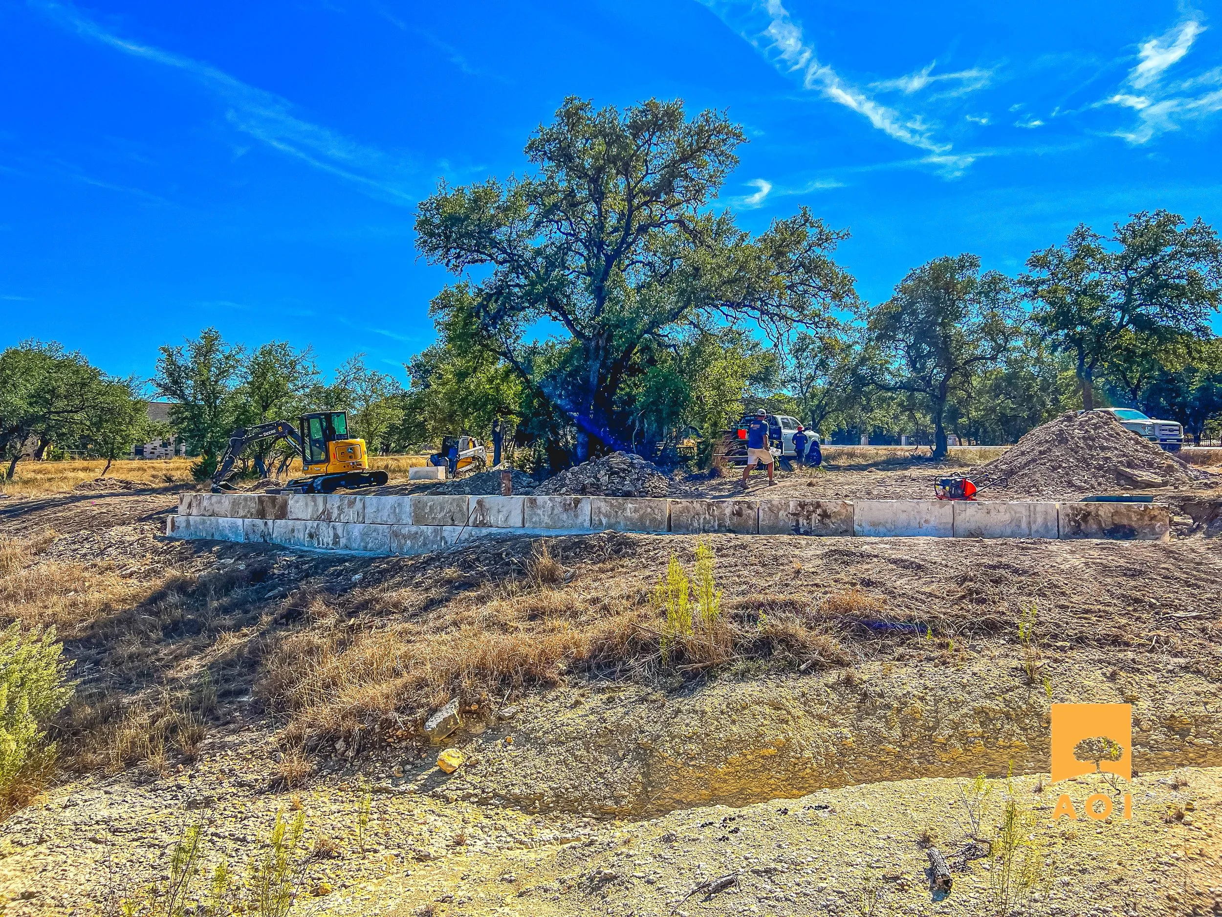 Construction workers and equipment building a foundation in a rural area with trees and a blue sky.