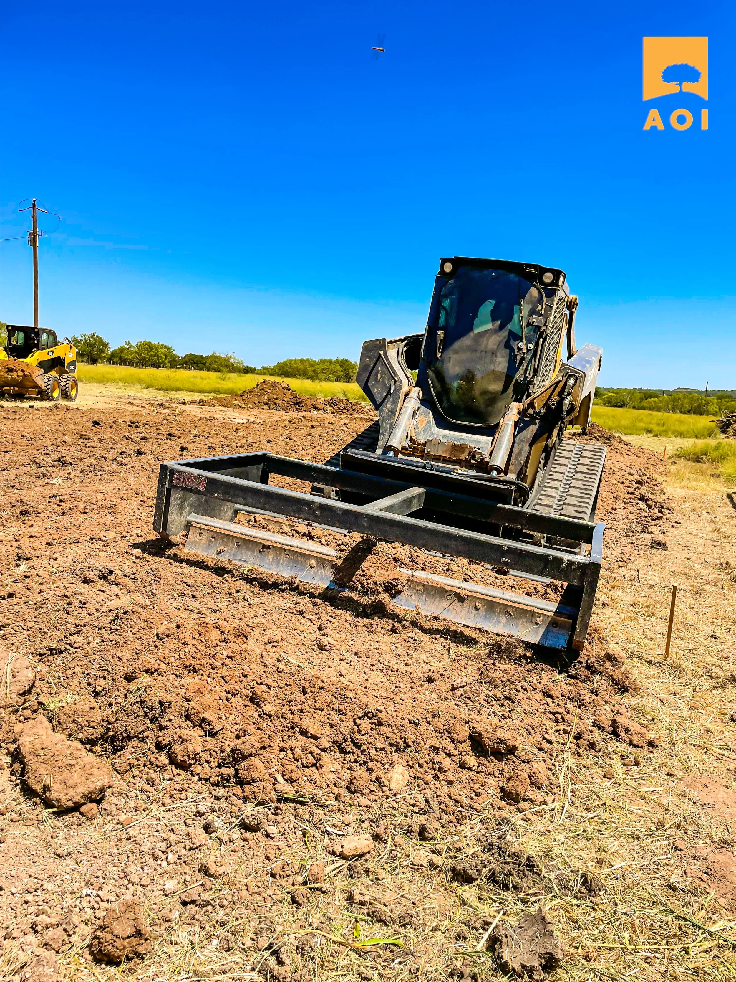 A construction site with a bulldozer on dirt, an excavator in the background, and a clear blue sky.