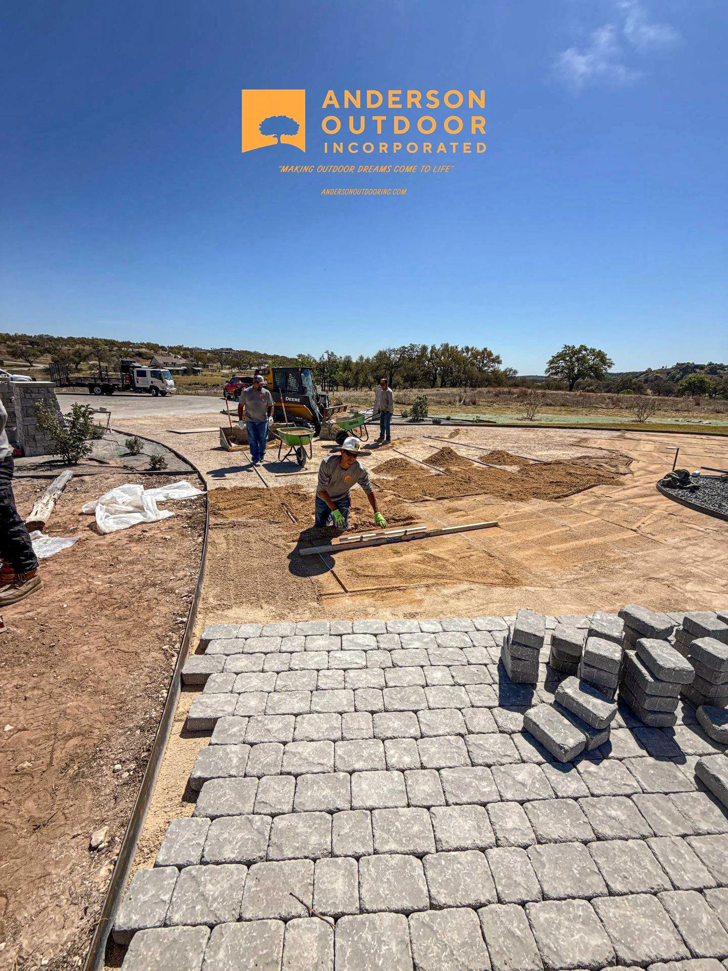 Construction workers laying paving stones on a pathway in an outdoor landscape under clear blue sky. There are stacks of stones in the foreground and a construction vehicle in the background with open land and trees.