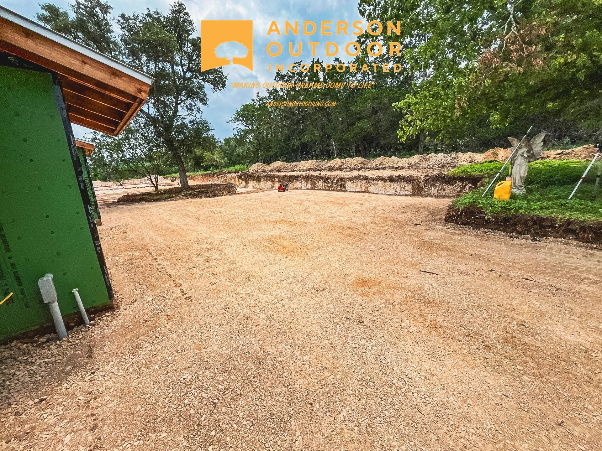 Construction site with cleared and leveled dirt area, partially constructed building with green sheathing on the left, trees and tools on the right, and orange safety equipment in the background. Overlay logo for Anderson Outdoor Incorporated.