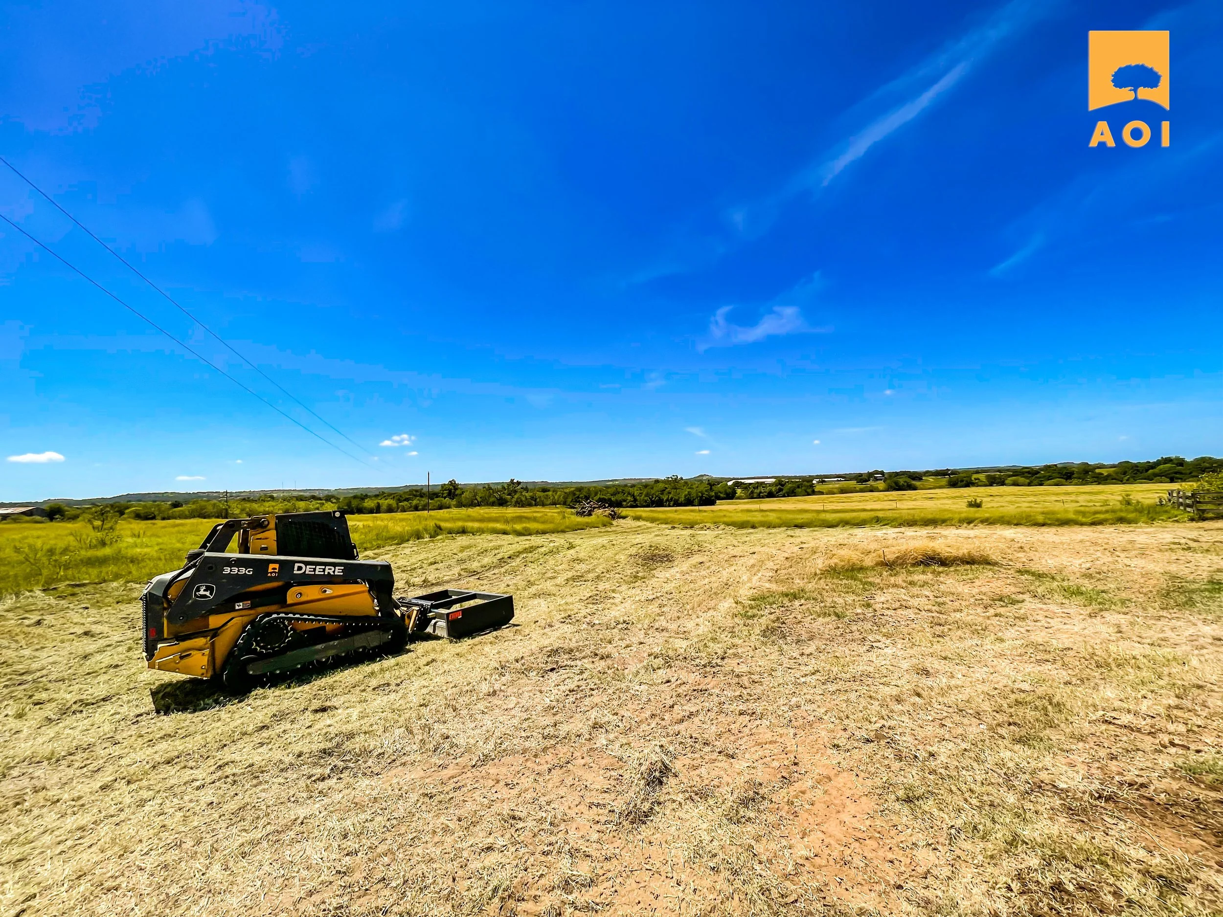 A John Deere mini excavator on a freshly mowed field under a bright blue sky with a few scattered clouds.