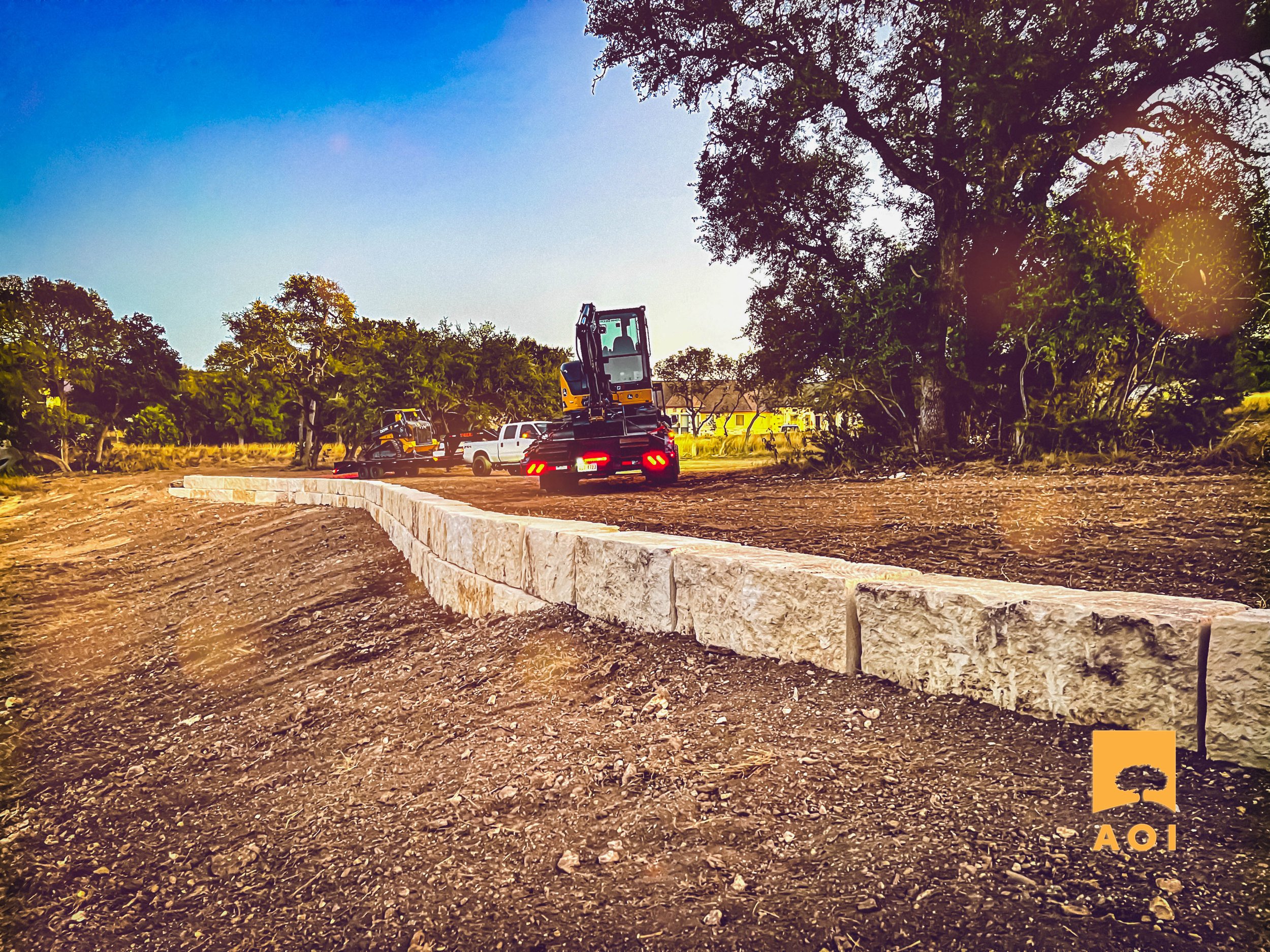 Construction site with a curved stone wall, construction vehicles, trucks, and machinery, surrounded by trees and a clear sky.