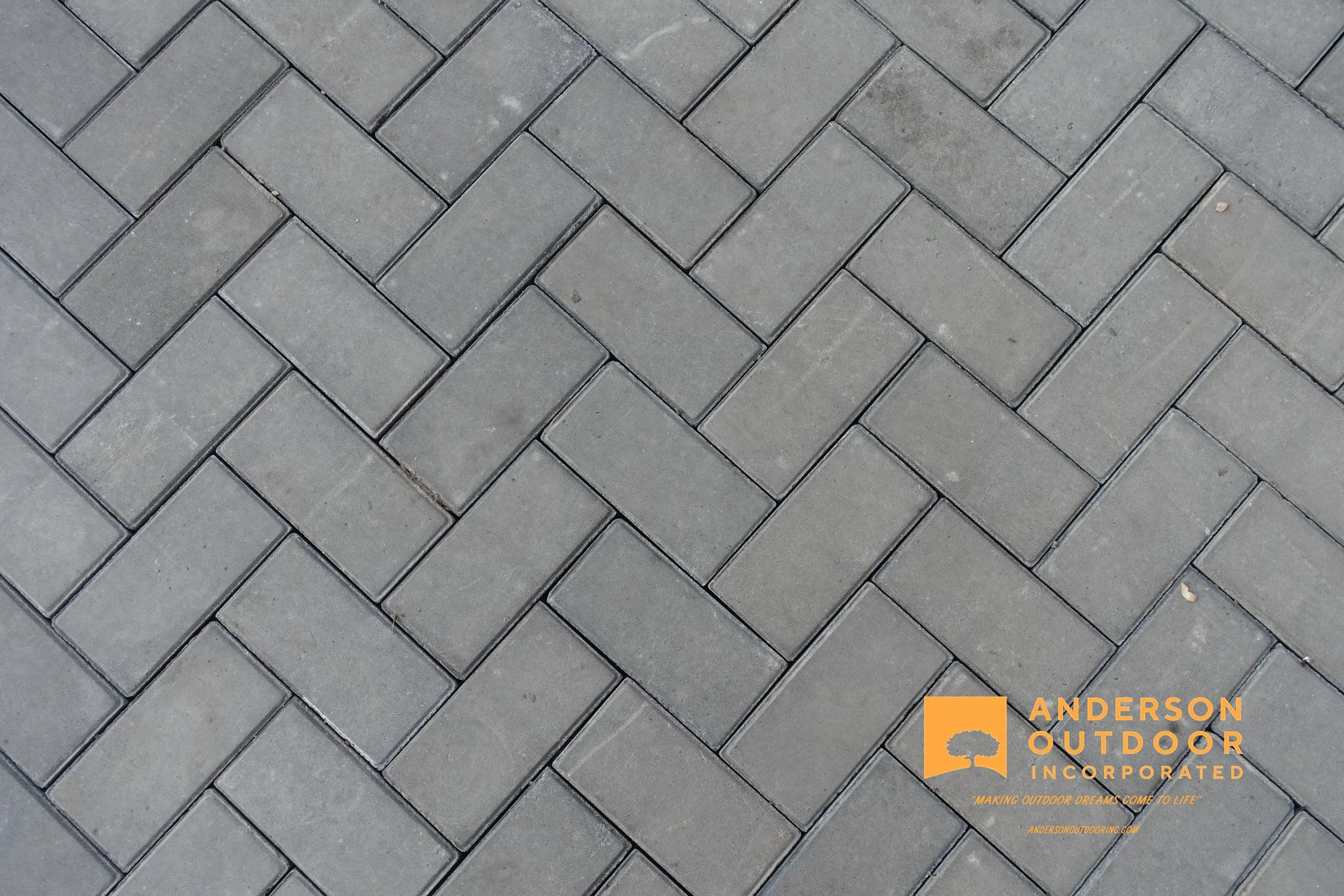 A close-up view of grey interlocking concrete pavers arranged in a herringbone pattern on a sidewalk or patio, with a logo of Anderson Outdoor Incorporated in the bottom right corner.