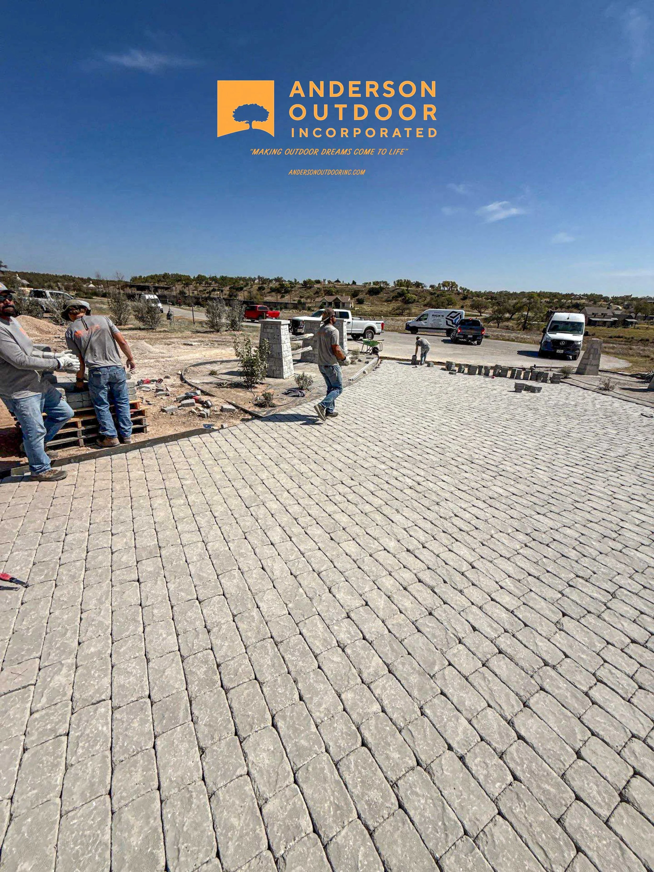 Construction workers laying brick pavers on a driveway under a clear blue sky, with trucks and a van parked in the background, and an advertisement for Anderson Outdoor Inc. at the top.