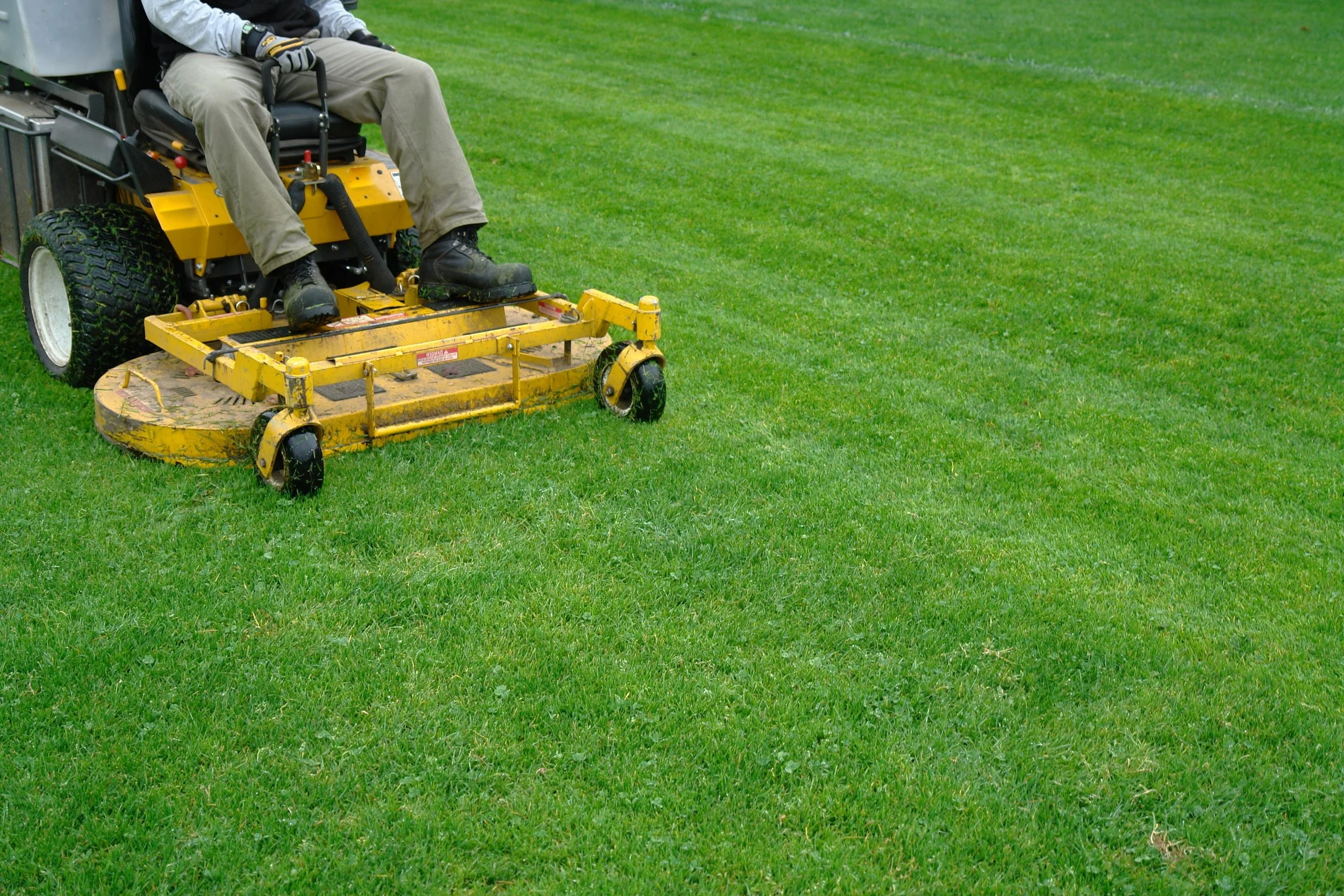 Close-up of a person operating a yellow riding lawn mower cutting green grass in a yard.