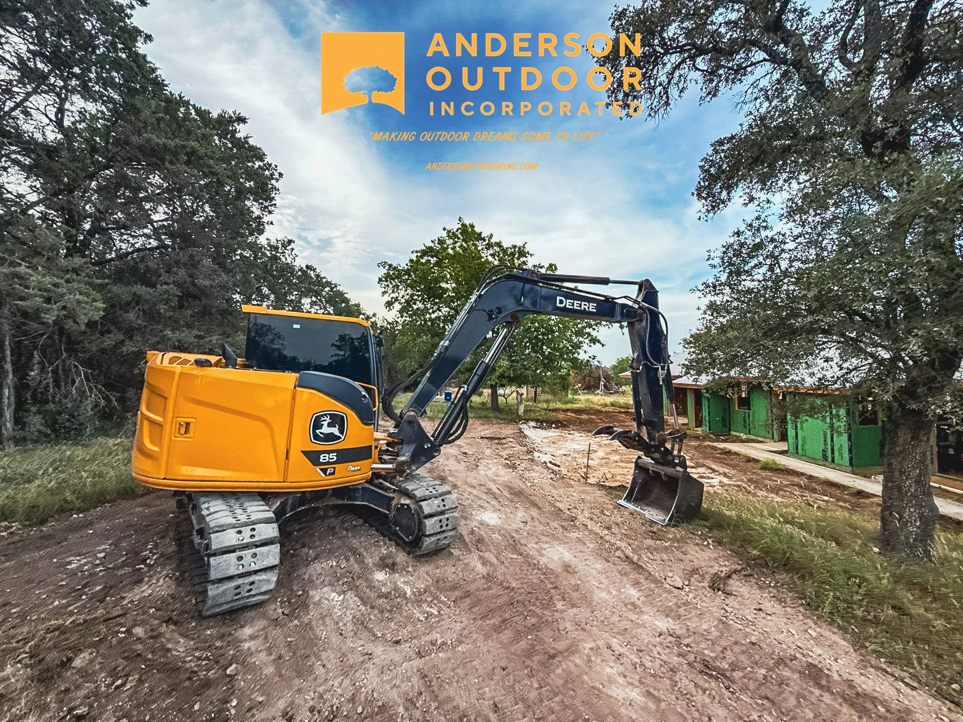 A yellow and black John Deere mini excavator is parked on a dirt trail in an outdoor setting with trees and green buildings nearby.