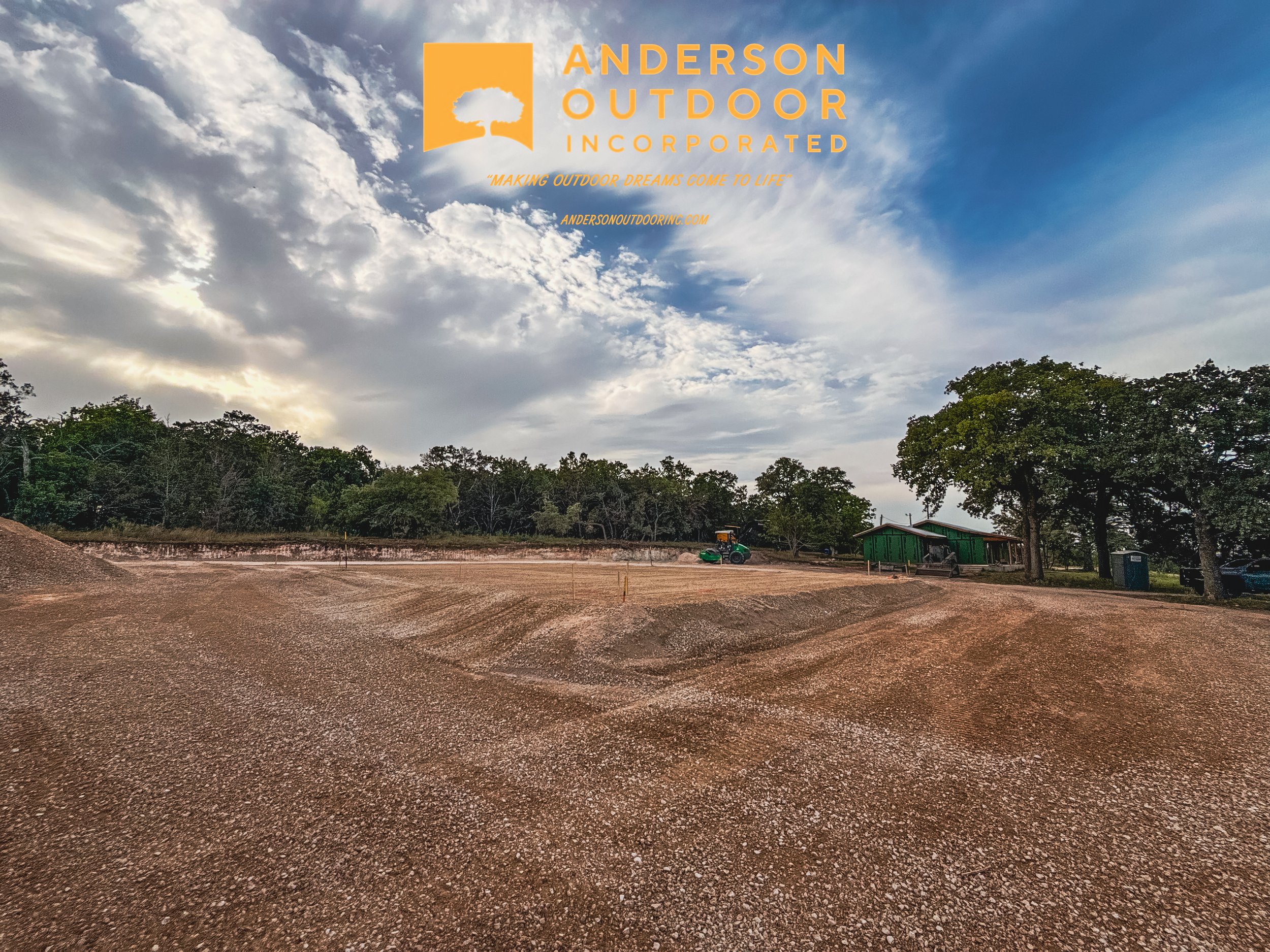 Construction site with a large dirt area, a green building, a tractor, and trees in the background under a cloudy sky, with logo and text for Anderson Outdoor Incorporated in the upper left corner.