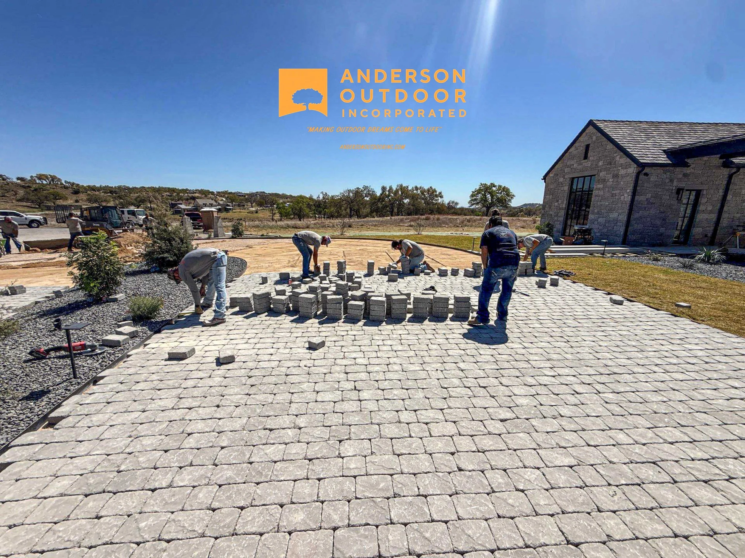 Workers installing a stone patio in a backyard near a house, with construction equipment and a blue sky.