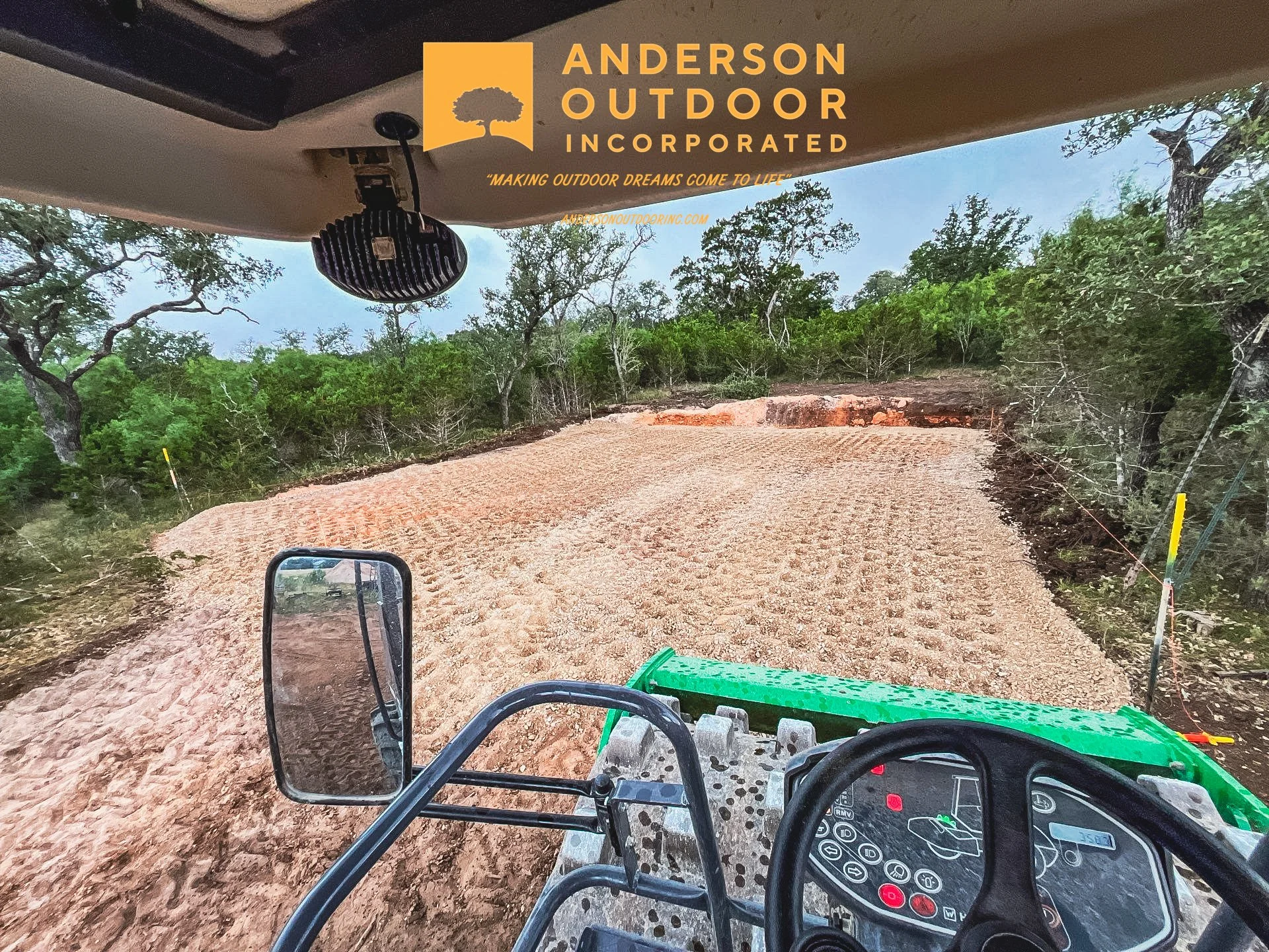 View from inside a construction vehicle on a dirt clearing surrounded by trees with a yellow sign and logo for Anderson Outdoor Incorporated at the top of the image.