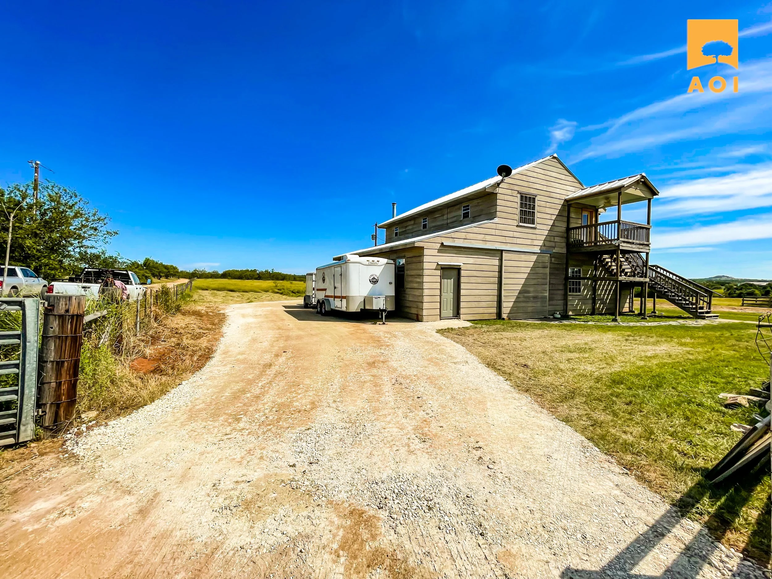 A rural property featuring a two-story wooden house with an exterior staircase, a gravel driveway, a white trailer, and parked vehicles, under a clear blue sky.