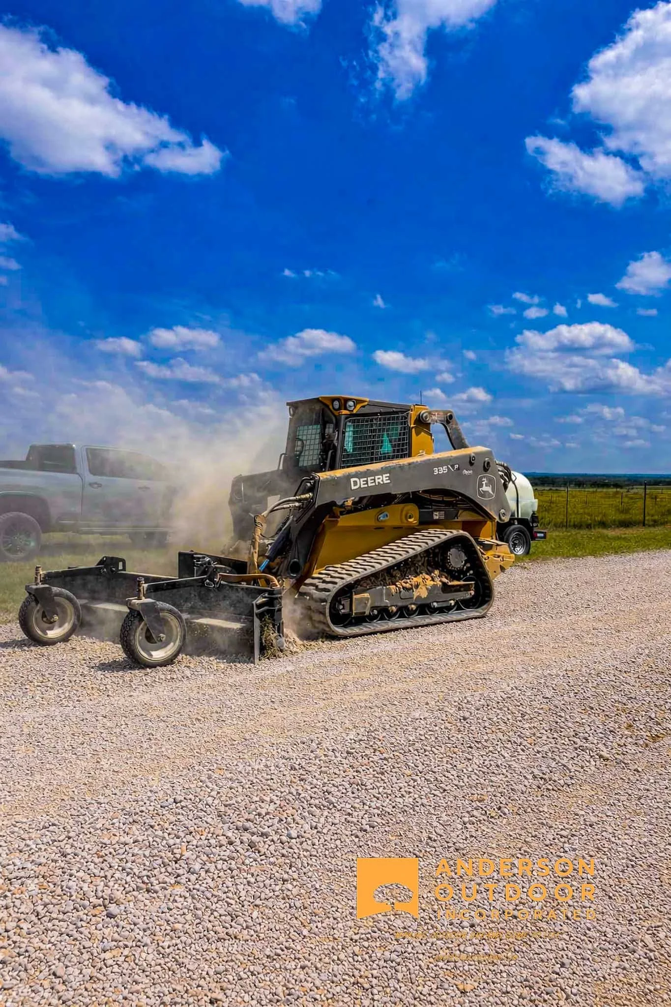 A large yellow road grader operating on a construction site during sunset, with dust and gravel being moved, and a colorful sky with clouds in shades of orange and blue.