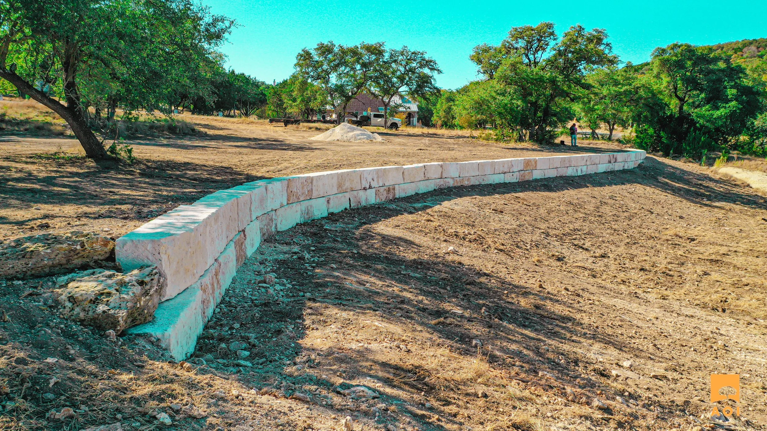 A curved stone retaining wall on a construction site with dirt and grass. In the background, there are trees, a house, a person, and vehicles under a clear blue sky.