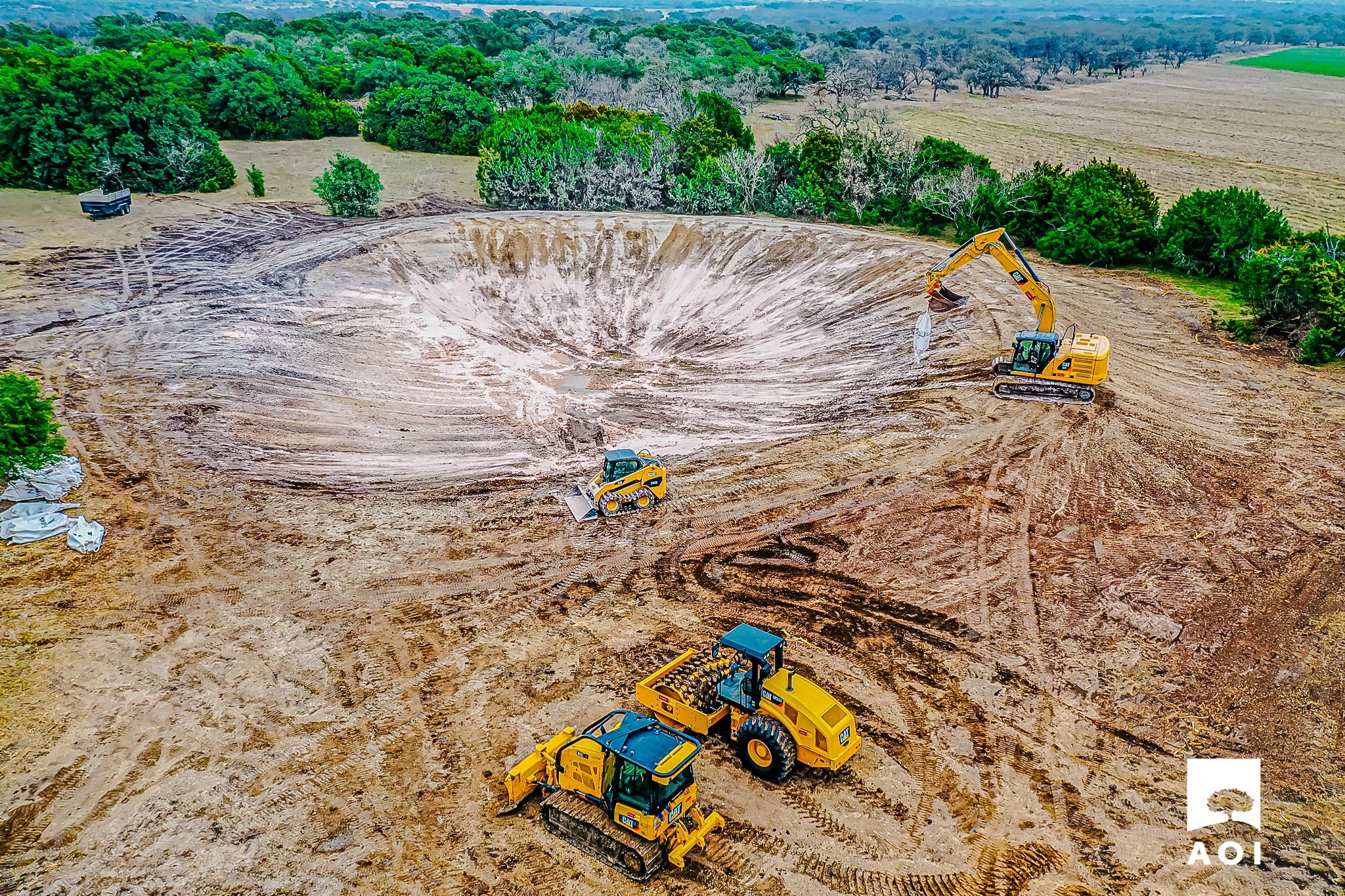 Construction site with heavy machinery, including excavators and bulldozers, shaping a large earth pit in a rural area surrounded by trees and open fields.