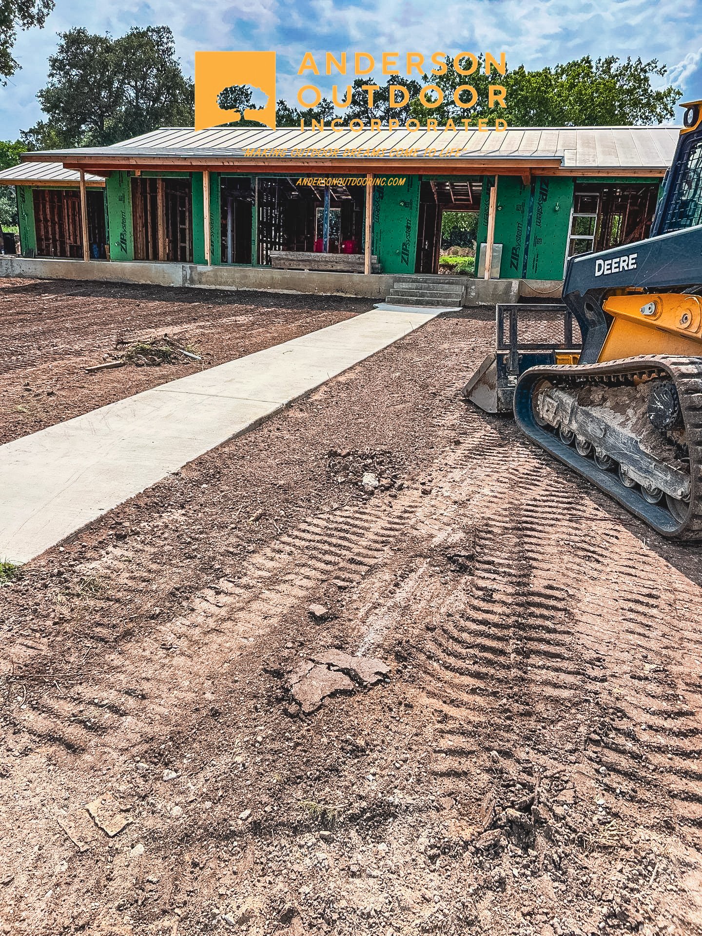 Construction site with a concrete walkway leading to a partially built structure with green sheathing, surrounded by dirt, and a small excavator on the right.