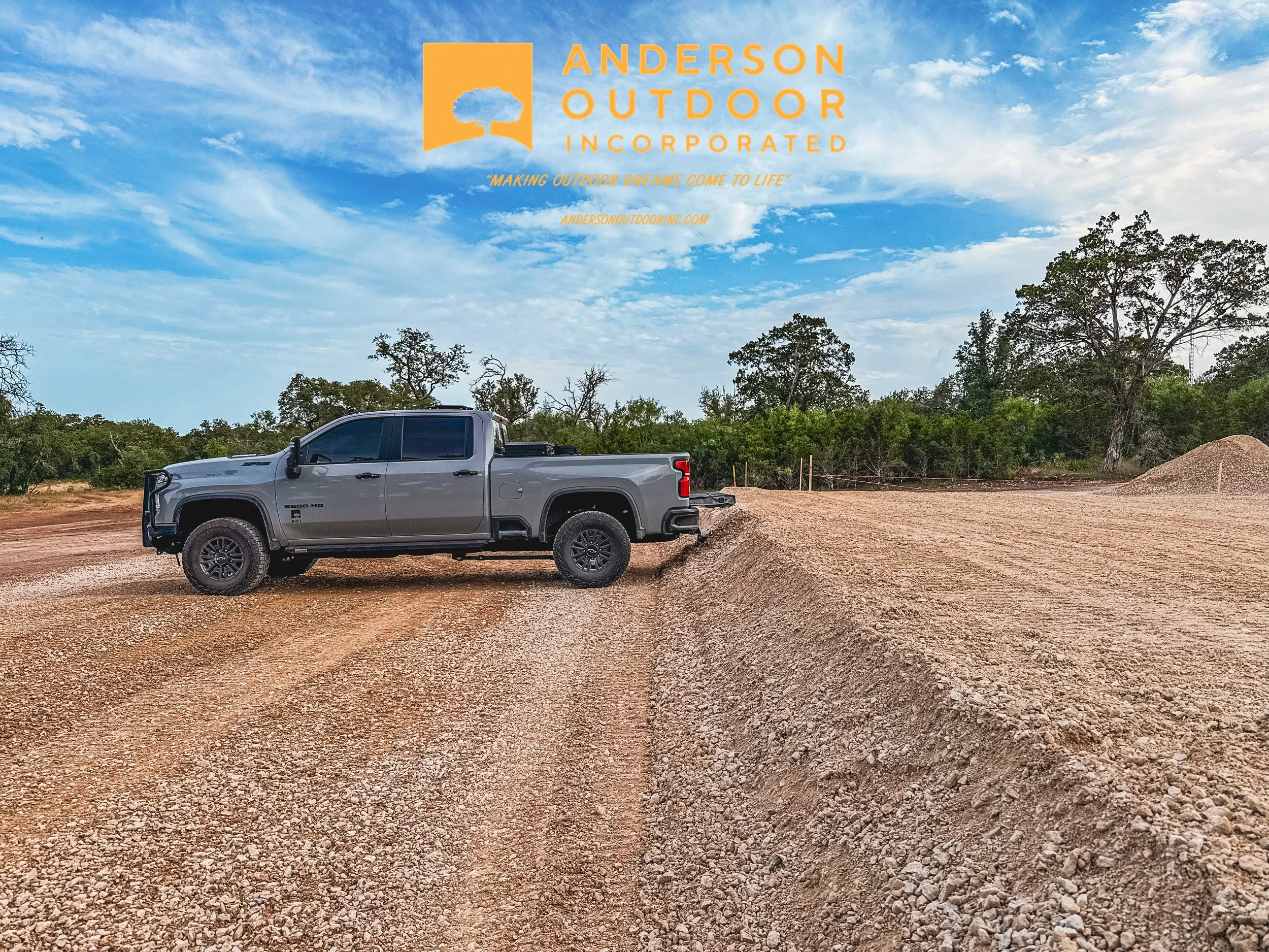A gray pickup truck parked on a gravel lot under a blue sky with scattered clouds, with trees in the background and a mound of gravel to the right, featuring overlay text of an orange logo and company name Anderson Outdoor Incorporated.