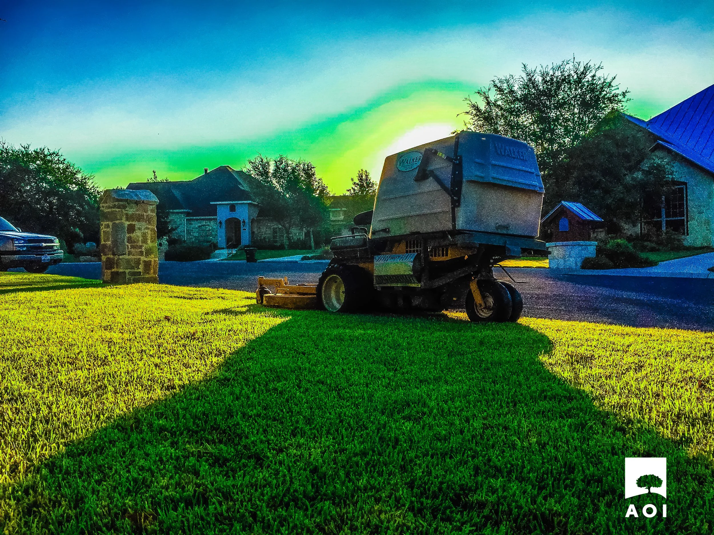A lawn mower is on a freshly cut grass patch in a suburban neighborhood during sunset or sunrise, with houses and trees in the background.