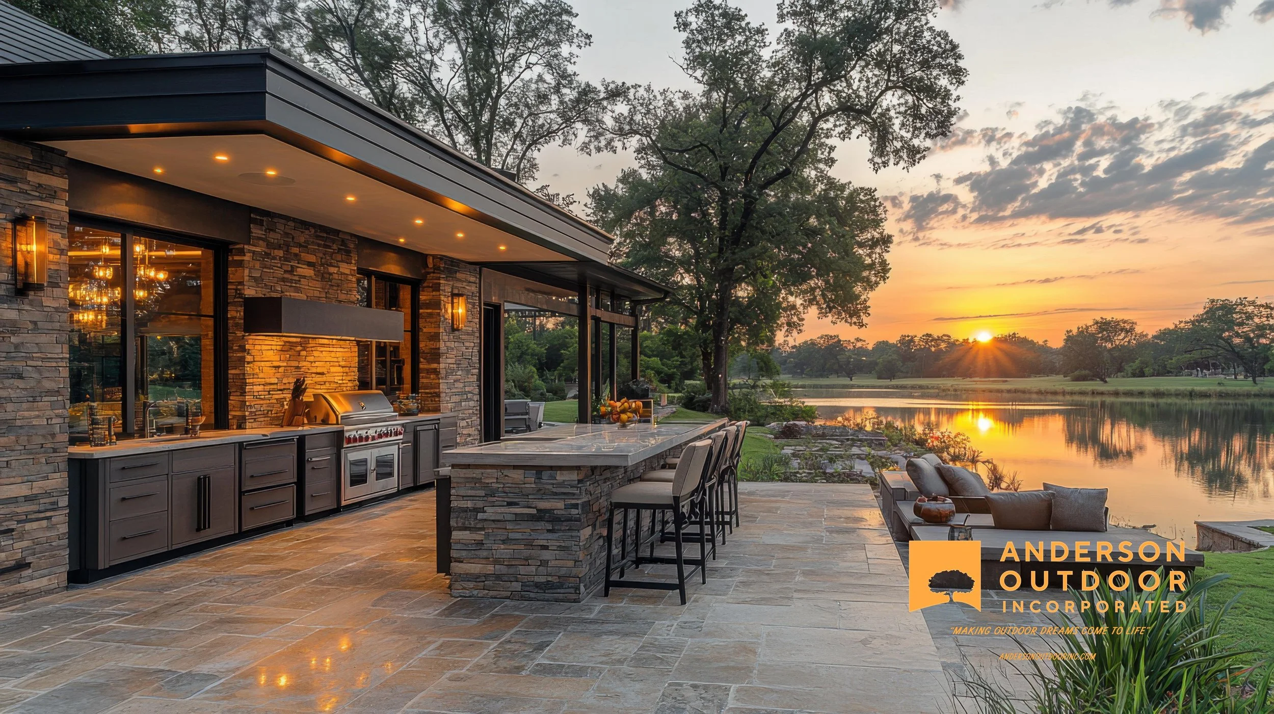 Outdoor kitchen and patio area with a built-in grill, bar seating, and a view of a lake and trees during sunset.