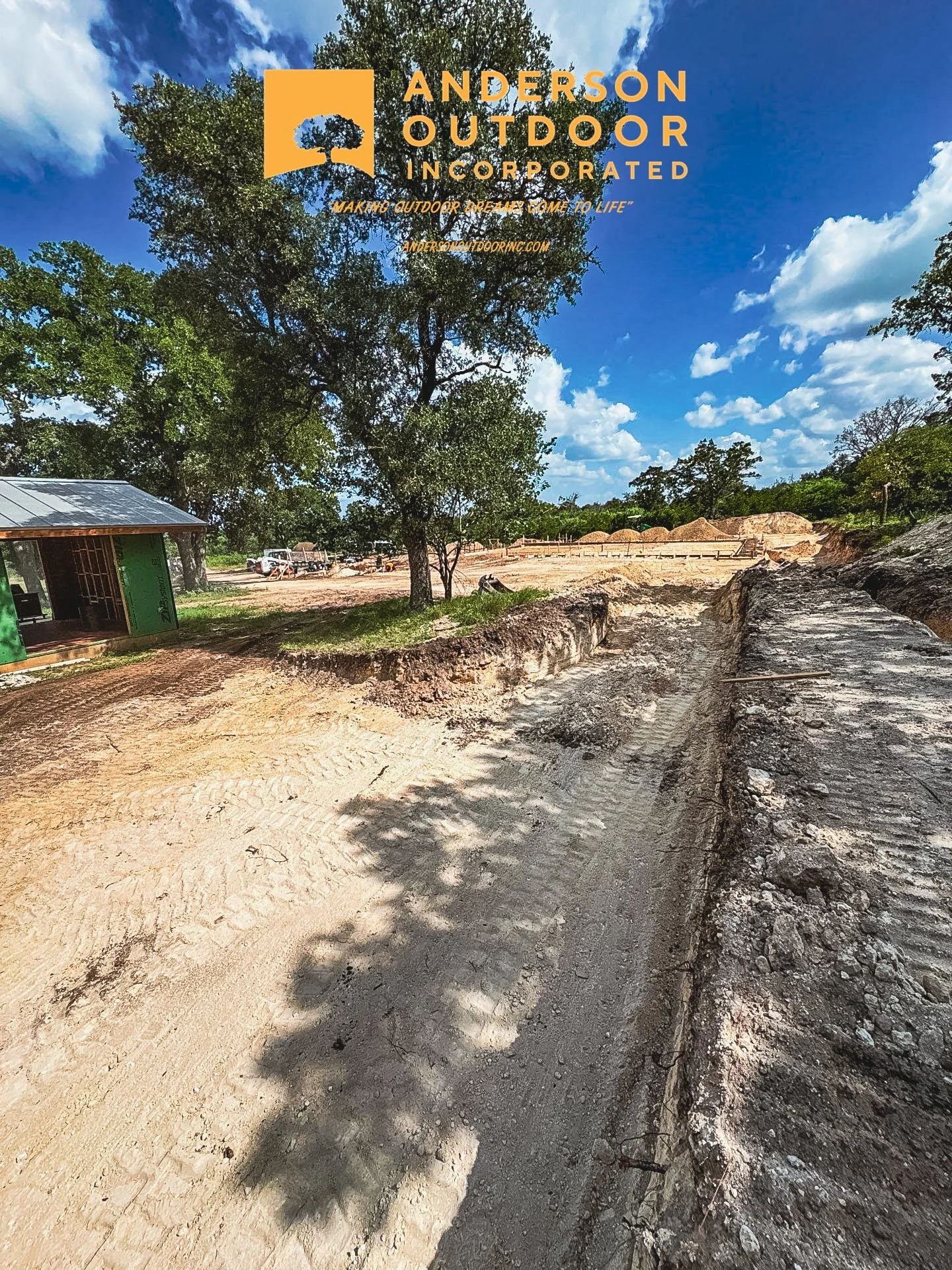 Construction site with dirt road, trees, and blue sky with clouds, logo of Anderson Outdoor Incorporated at the top.