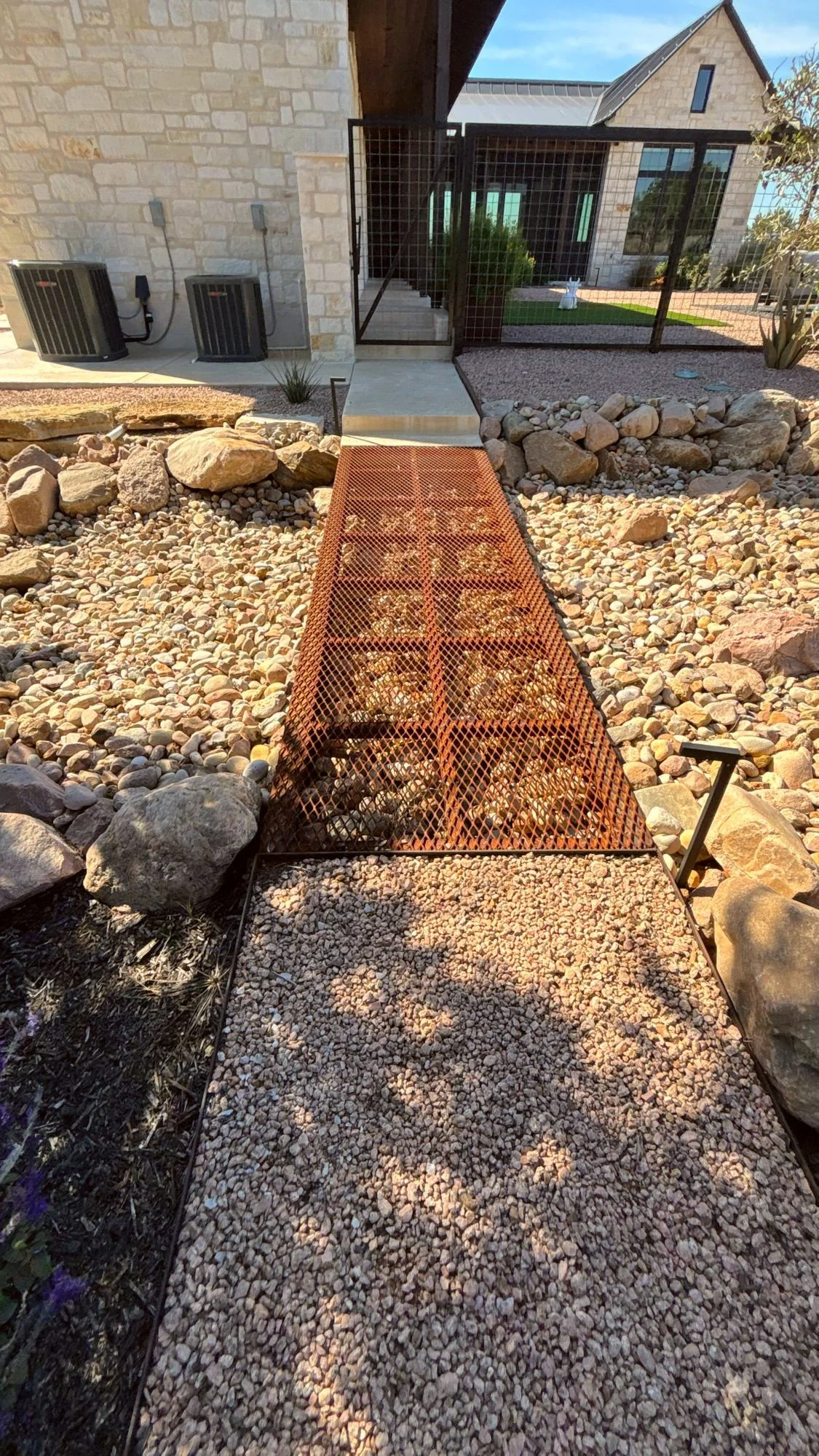 Steel grate footbridge crossing a river rock dry creek bed beside a limestone home in the Texas Hill Country.