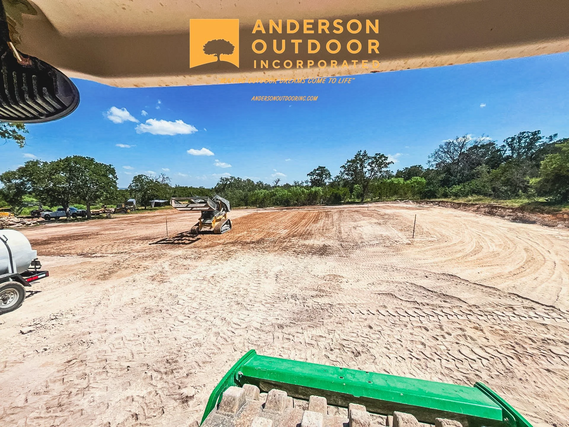 A construction site with dirt ground, a small excavator, and cars parked in the background. Trees and a blue sky with scattered clouds are visible. The image is taken from inside a construction vehicle.