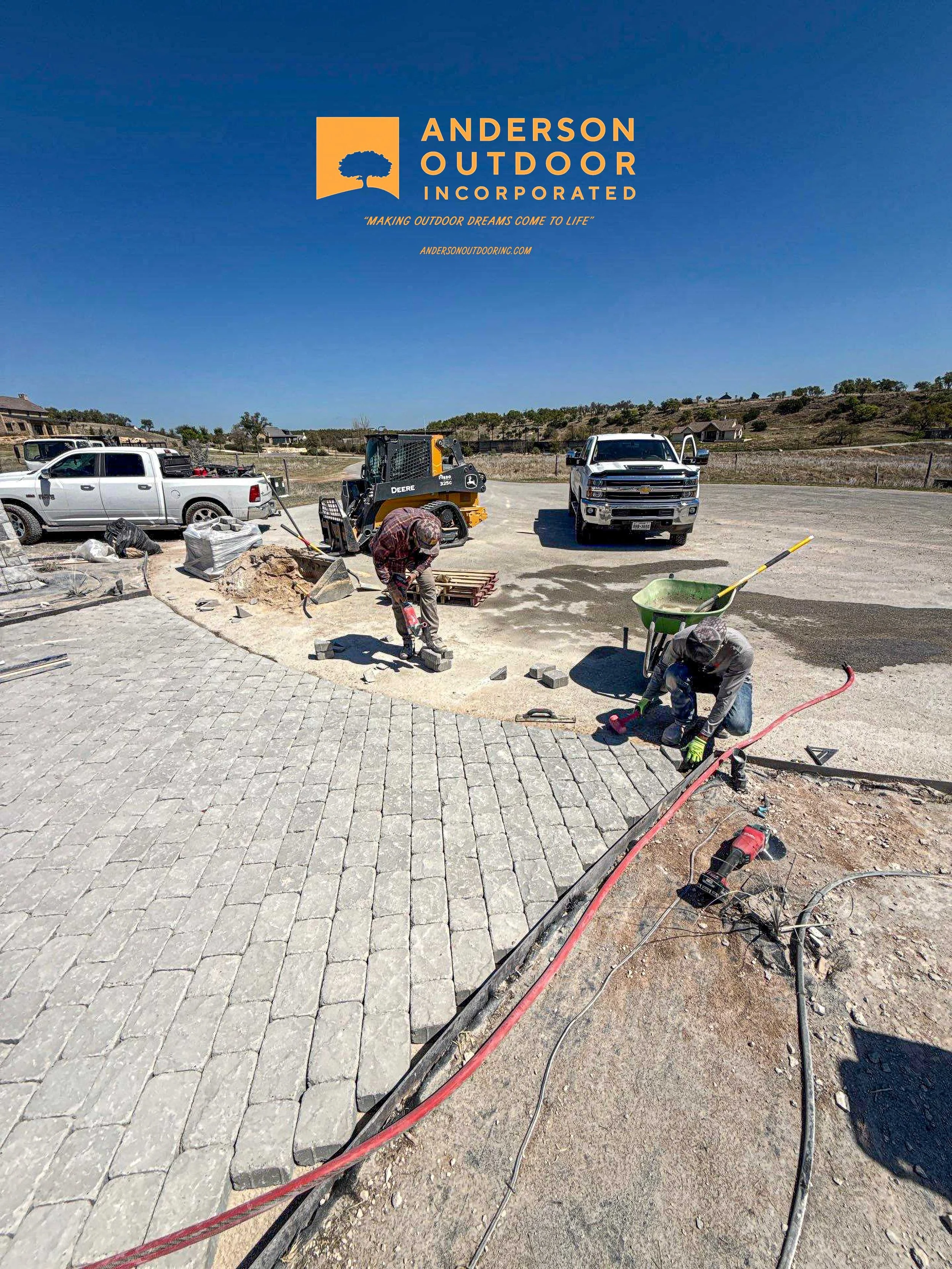 Workers installing pavers on a sidewalk at a construction site, with trucks and construction equipment nearby under a clear blue sky.