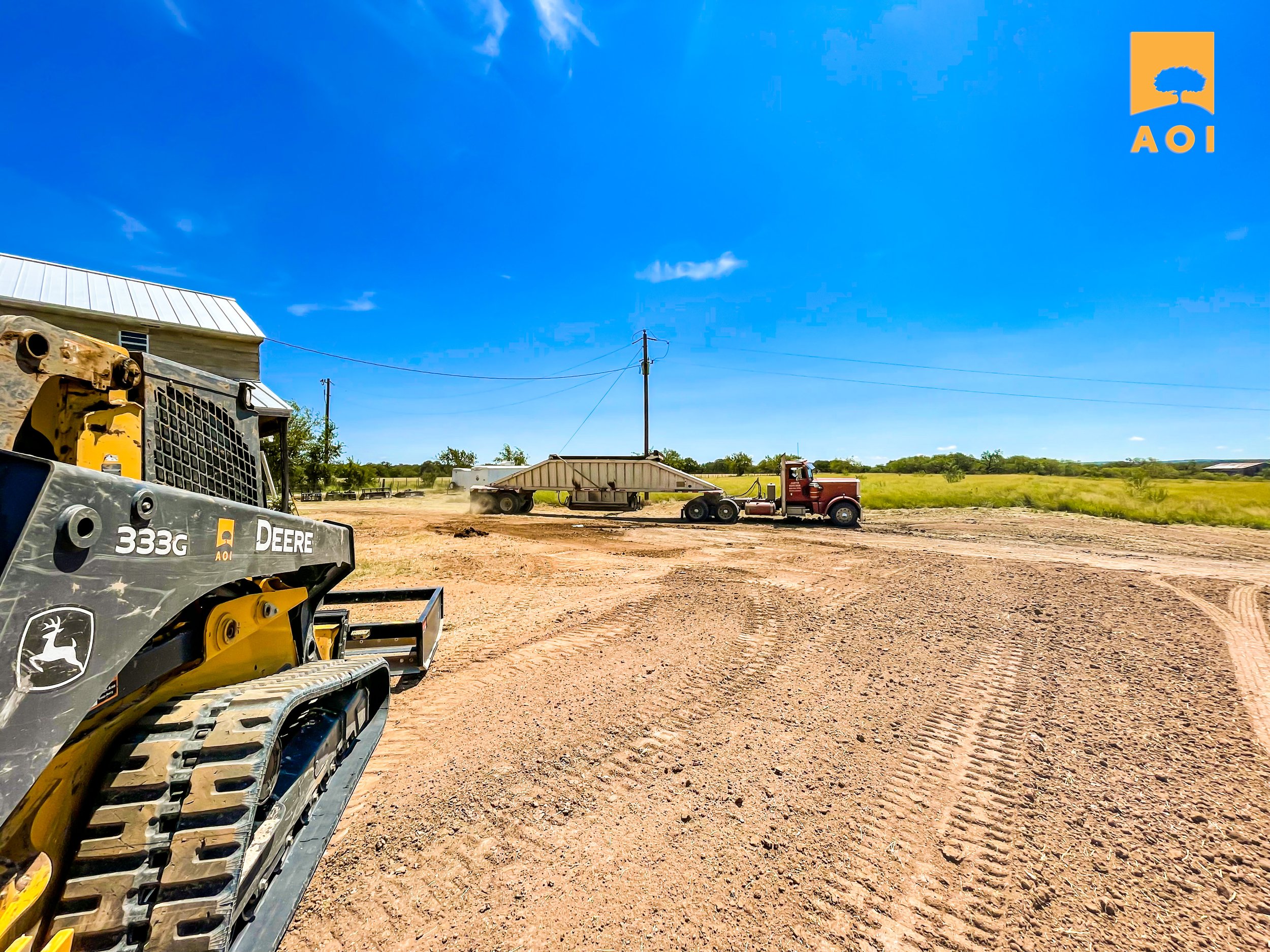 A construction site with a John Deere skid steer loader in the foreground on the left, a large red truck with a trailer in the middle, and a vast green field with a clear blue sky in the background.