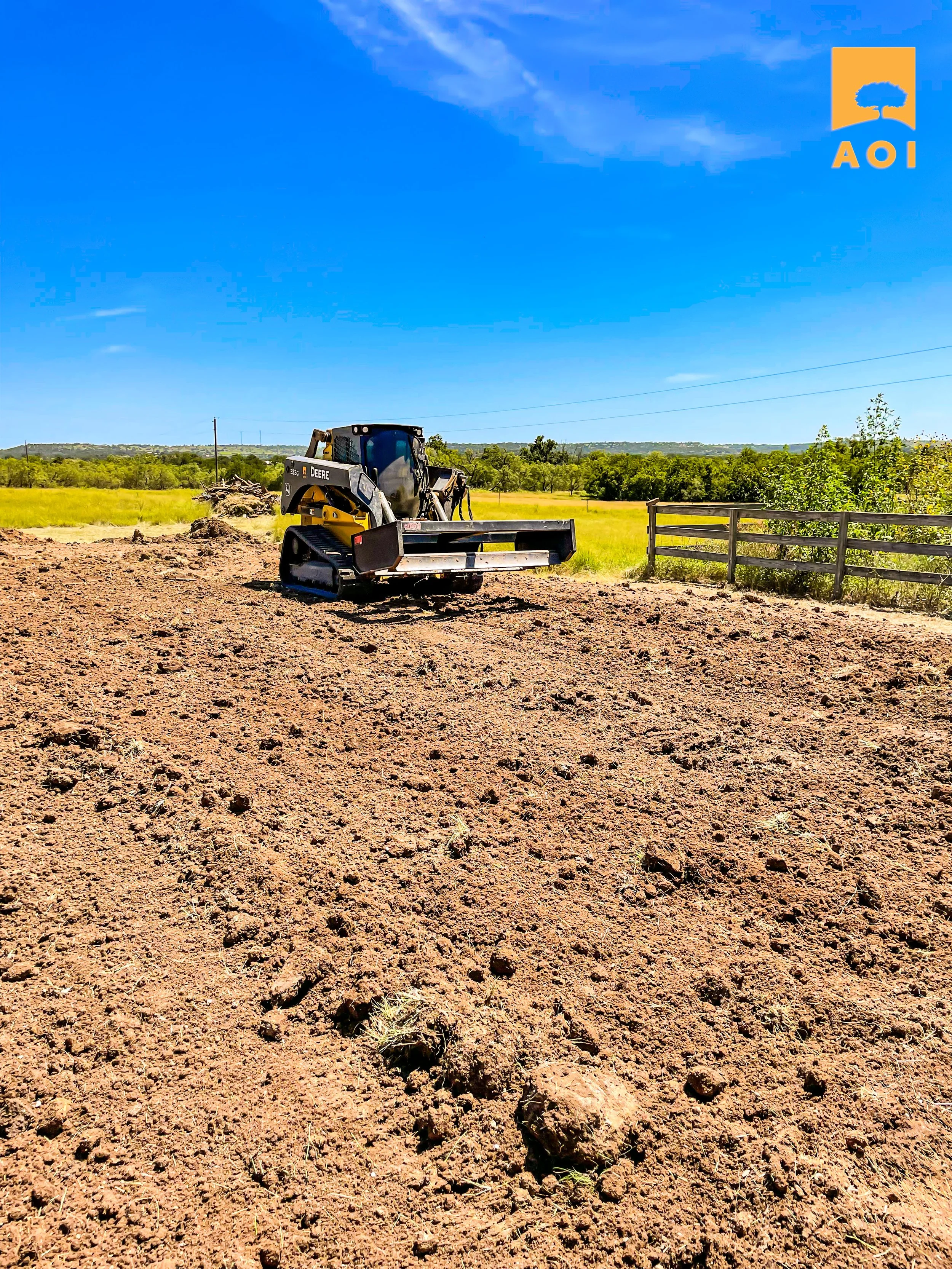 A John Deere bulldozer on a dirt plot of land with a grassy field and trees in the background under a bright blue sky. The AOI logo is in the top right corner.