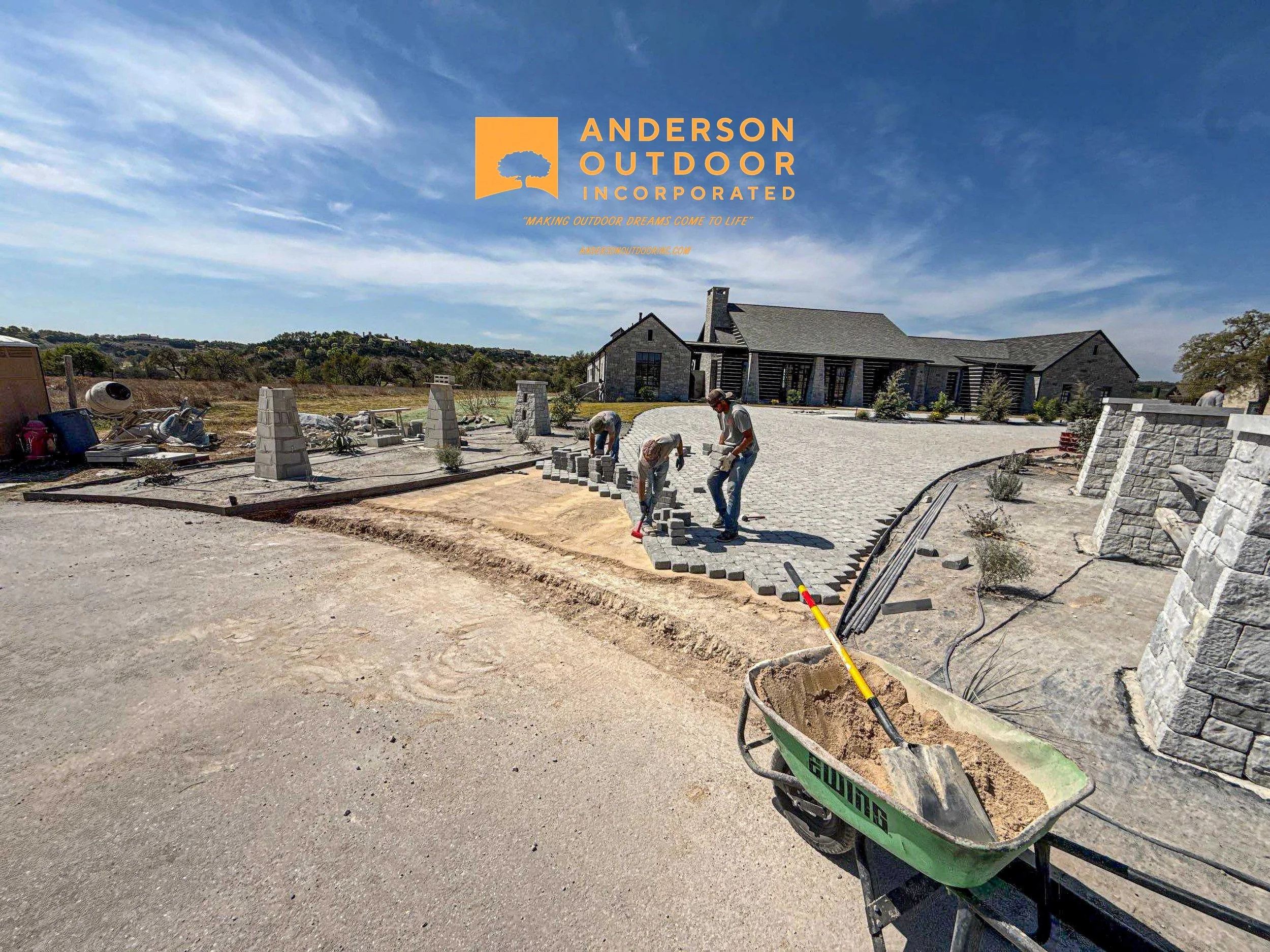 Construction workers installing paving stones in a driveway of a large house, with construction equipment and materials around.