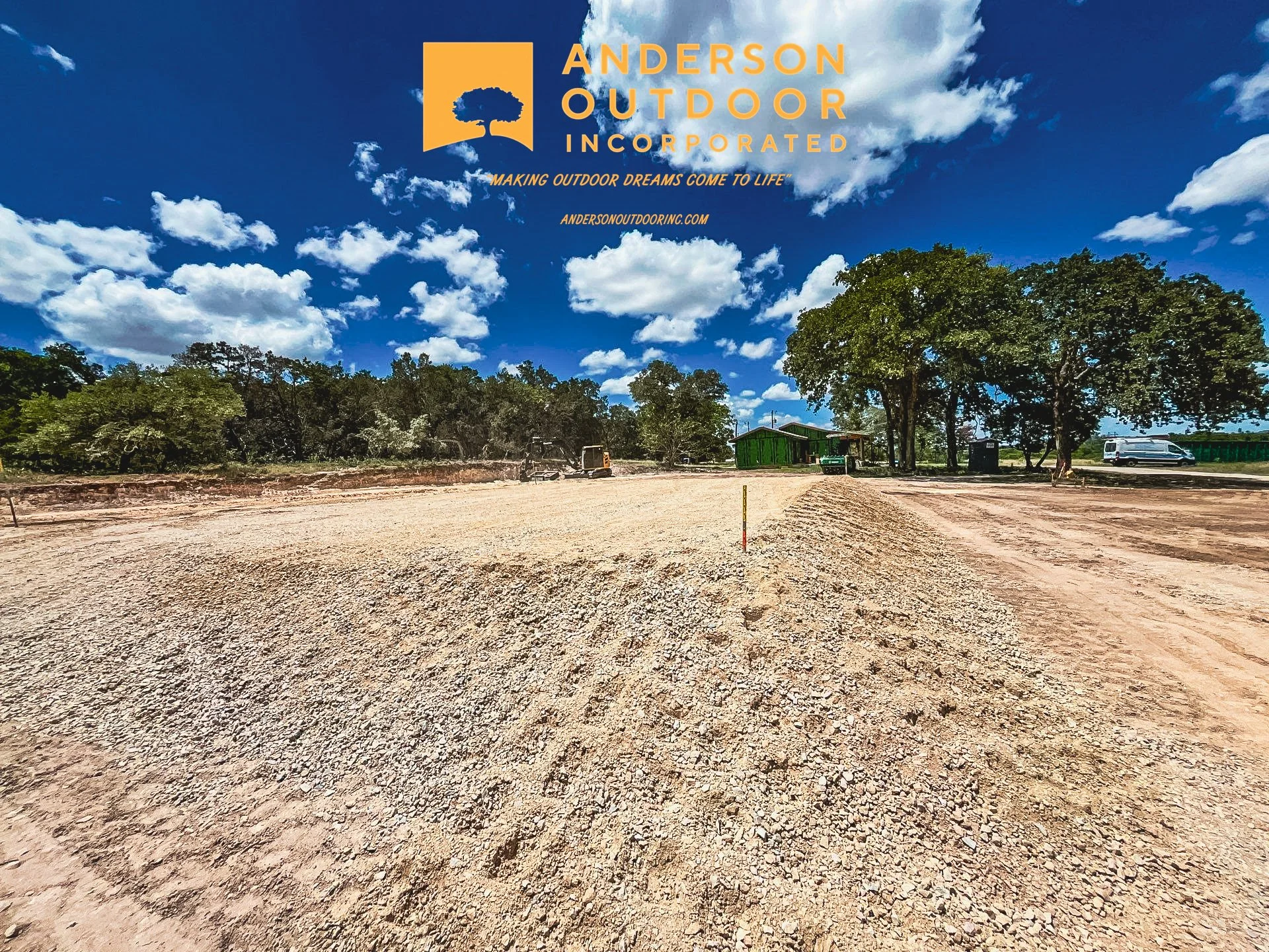 A construction site with a gravelly ground, scattered trees, and several buildings and vehicles in the background under a partly cloudy blue sky, with a company logo for Anderson Outdoor Inc. at the top.
