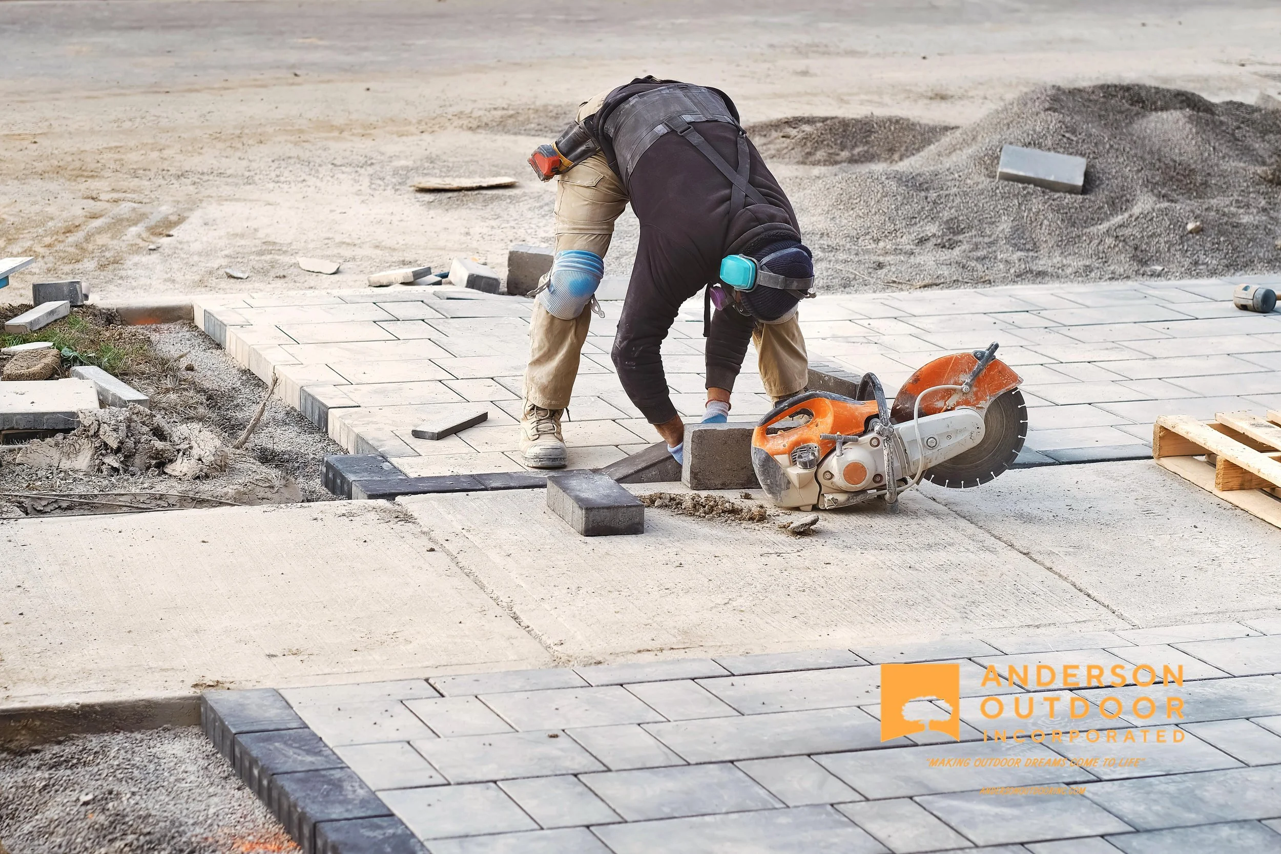 A worker installing paving stones on a sidewalk at a construction site. The worker is bending down, wearing protective gear, and using a power saw. There are sand piles and tools around, with a partially completed sidewalk area.
